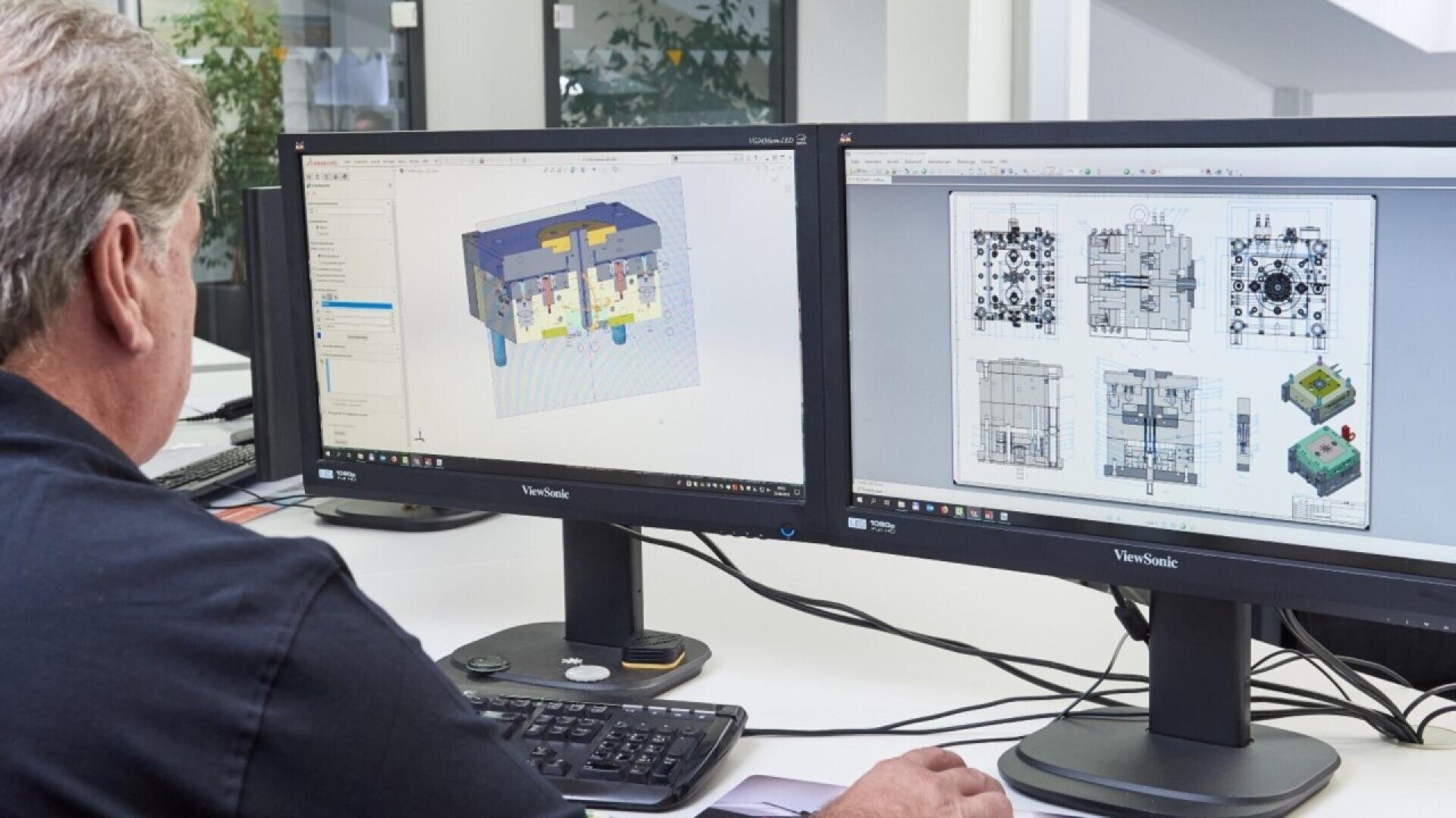 A person working at a desk is viewing 3D mechanical designs and technical drawings on two computer monitors, with a keyboard and mouse in front of them. The workspace is bright and modern.