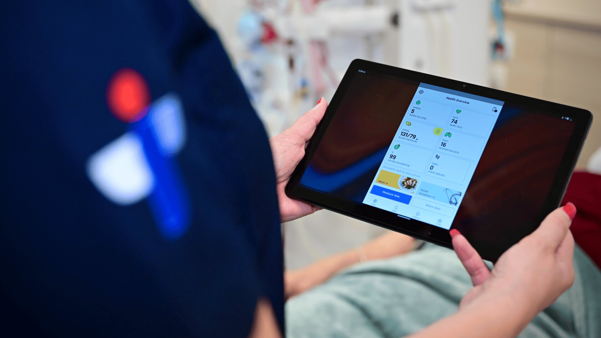 A person in medical scrubs holds a tablet displaying patient health data, while another individual lies in a hospital bed in the background.
