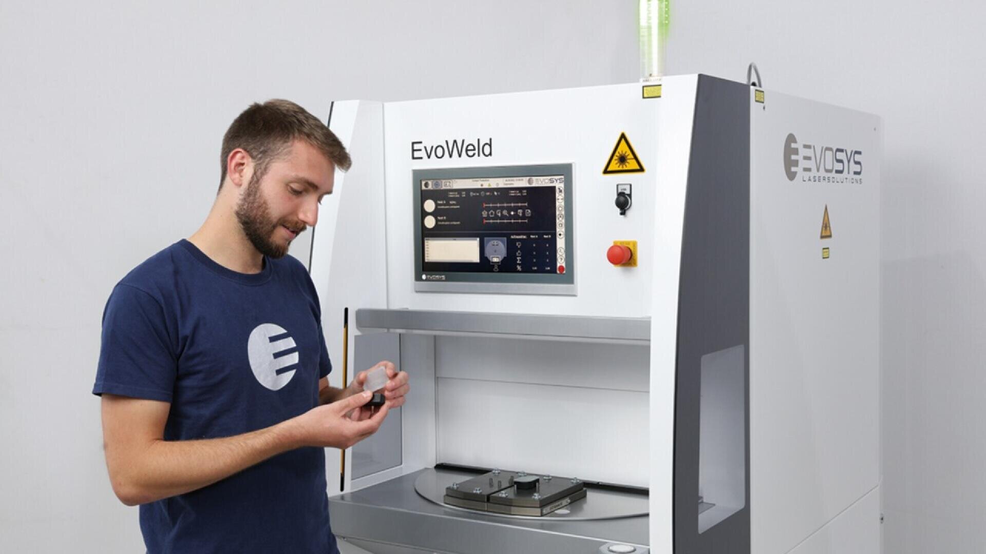 A man stands beside an EvoWeld machine, examining a small object in his hands. The machine has a control panel with buttons and a display screen, and he appears focused on his task.