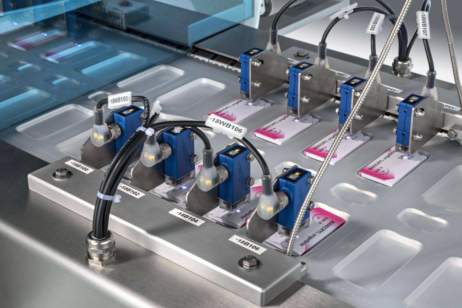 Nahaufnahme einer automatisierten Maschine mit Sensoren und beschrifteten Kabeln, die medizinische Testobjektträger auf einer Edelstahlplattform in einer Industrie- oder Laborumgebung prüft.