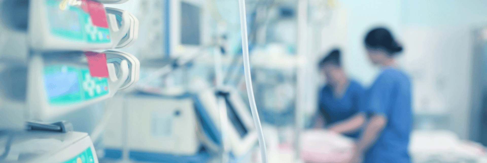Close-up of medical equipment and monitors in a hospital room, with two medical professionals in blue scrubs working in the blurred background. The environment appears clinical and sterile.
