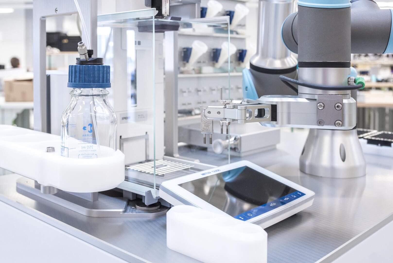 Ein Roboterarm arbeitet in einem Labor neben einer Glasflasche, einem digitalen Touchscreen-Display und verschiedenen Laborgeräten auf einer Metalloberfläche.