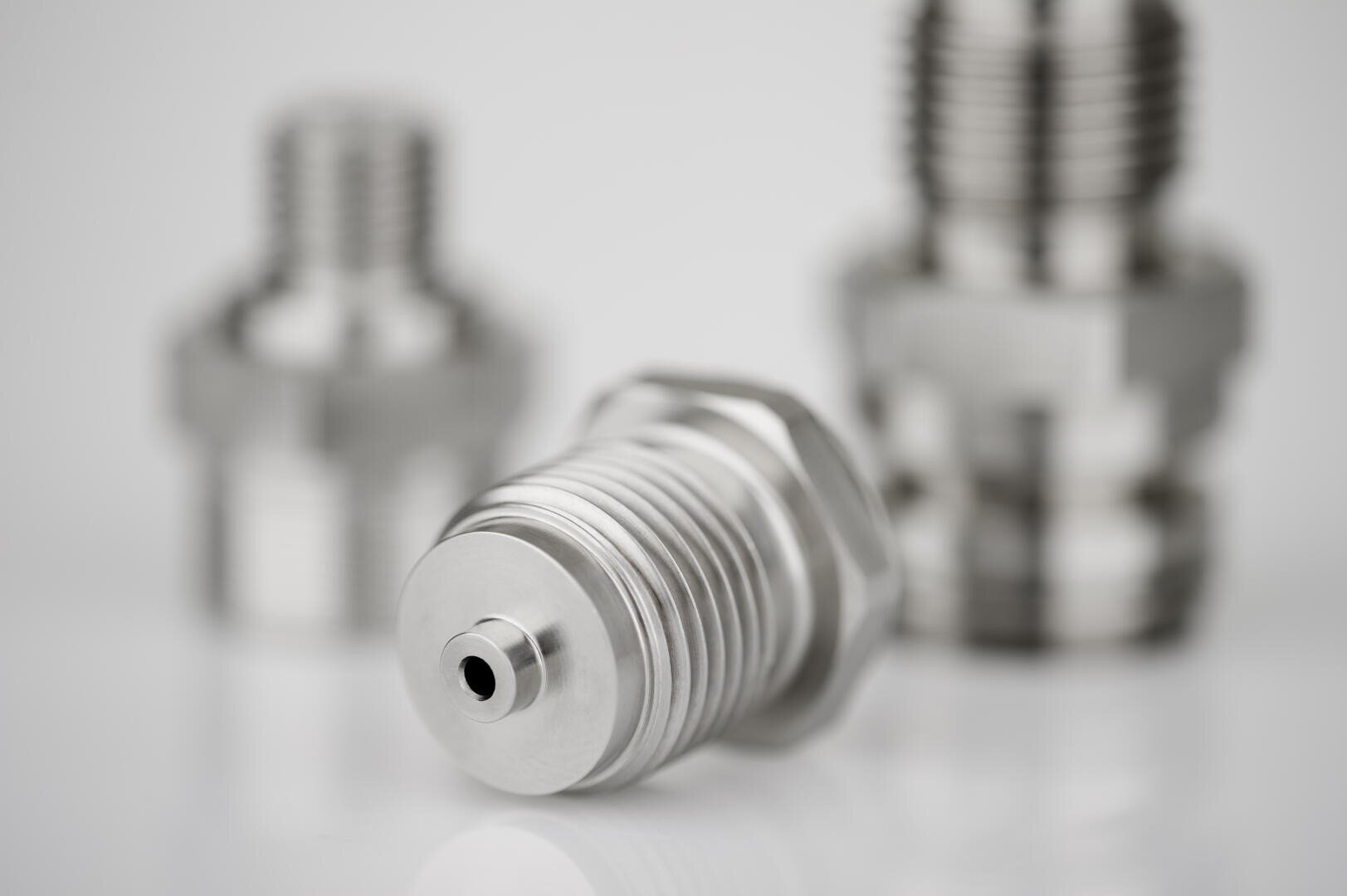 Close-up of three stainless steel industrial fittings, with one fitting in sharp focus laying horizontally in the foreground and two more blurred in the background on a white surface.