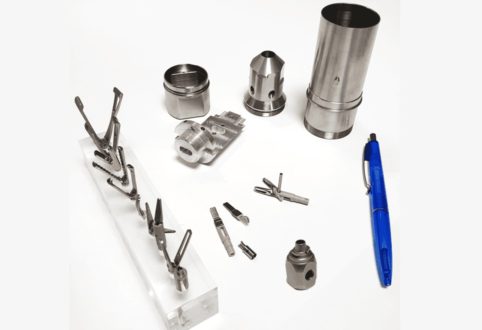 Several metal mechanical parts of varying shapes and sizes are arranged on a white surface alongside a blue pen for scale. Some parts are displayed on a transparent stand.