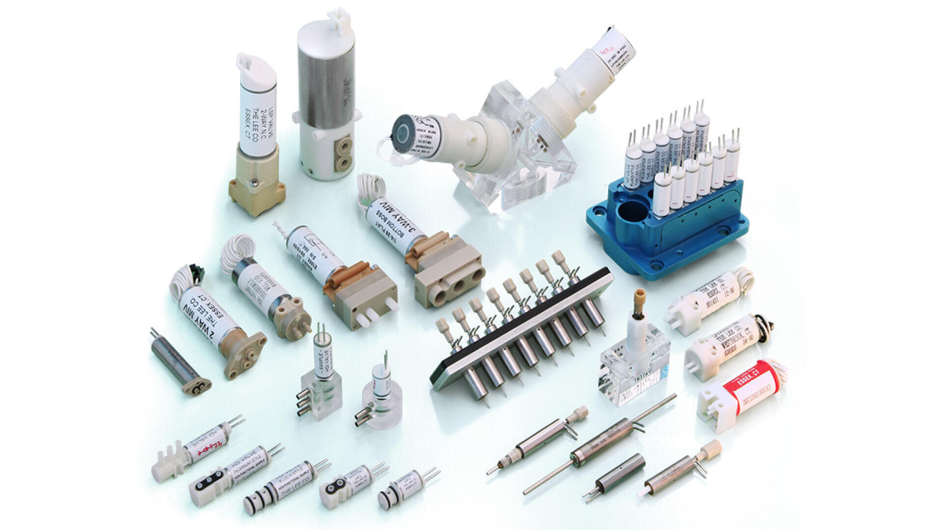 Eine Vielzahl von elektronischen und industriellen Bauteilen, darunter Sicherungen, Schutzschalter, Steckverbinder und Stecker, sind übersichtlich auf einer weißen Fläche angeordnet. Jedes Bauteil hat ein Etikett mit den technischen Daten.
