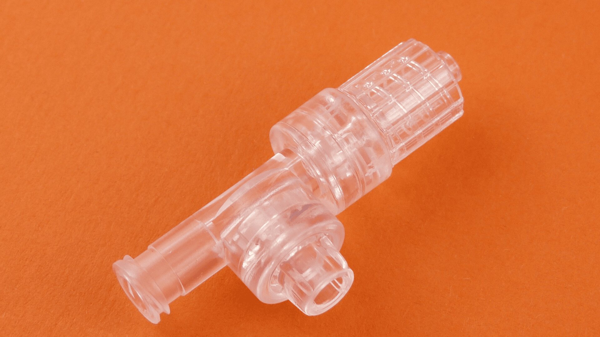 Auf orangefarbenem Hintergrund ist ein durchsichtiger medizinischer Kunststoffverbinder mit zylindrischen und geriffelten Abschnitten abgebildet.