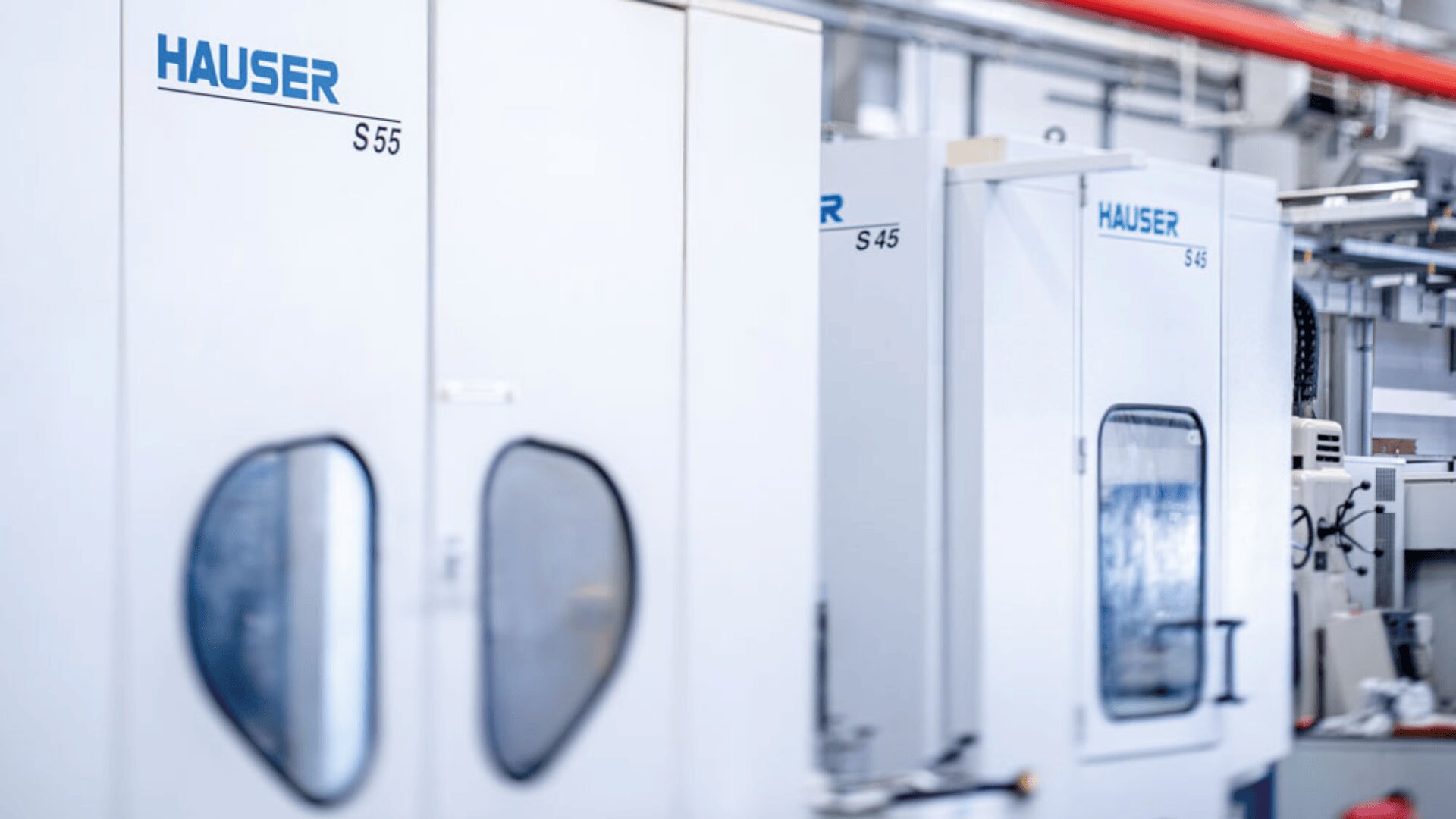 Mehrere weiße Industriemaschinen mit der Bezeichnung HAUSER, darunter die Modelle S55, S45 und S46, stehen in einer hellen, sauberen Fabrik- oder Produktionsumgebung aufgereiht.
