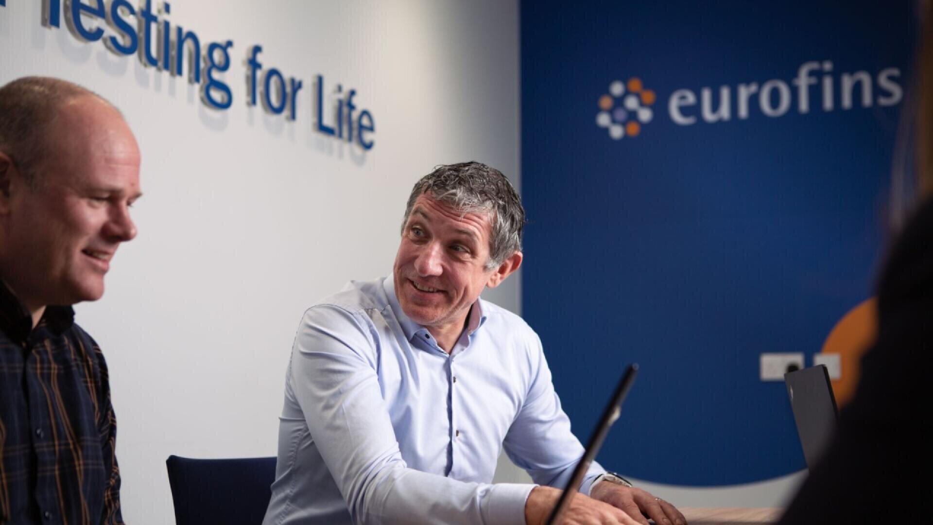 Two men sit at a table in an office with Testing for Life and eurofins visible on the wall behind them, engaged in conversation, with laptops in front of them.