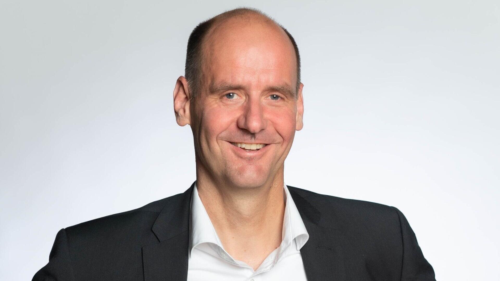A smiling man with a bald head wearing a dark suit jacket and white dress shirt stands against a plain, light background.