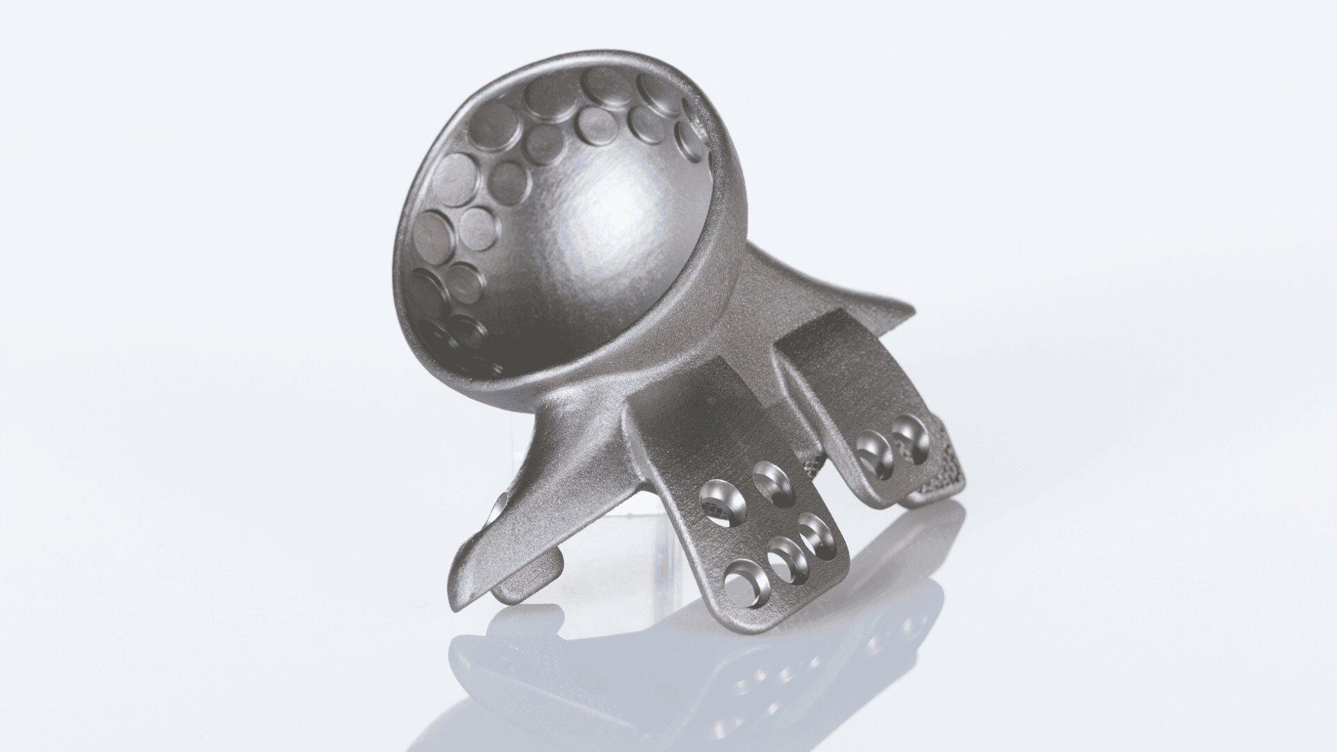 Ein metallisches, 3D-gedrucktes Objekt mit einer abgerundeten, konkaven Oberseite und mehreren flachen, perforierten Verlängerungen, platziert auf einer weißen Oberfläche.