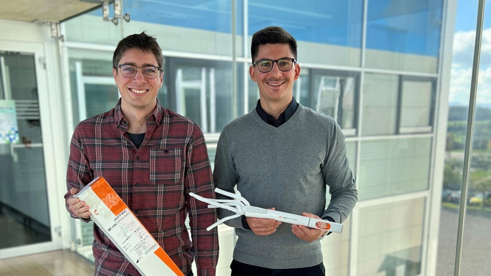 Two men stand indoors by large windows, both smiling. One holds a rectangular box, and the other holds a white foldable object. They are casually dressed and appear to be in a modern office or building.