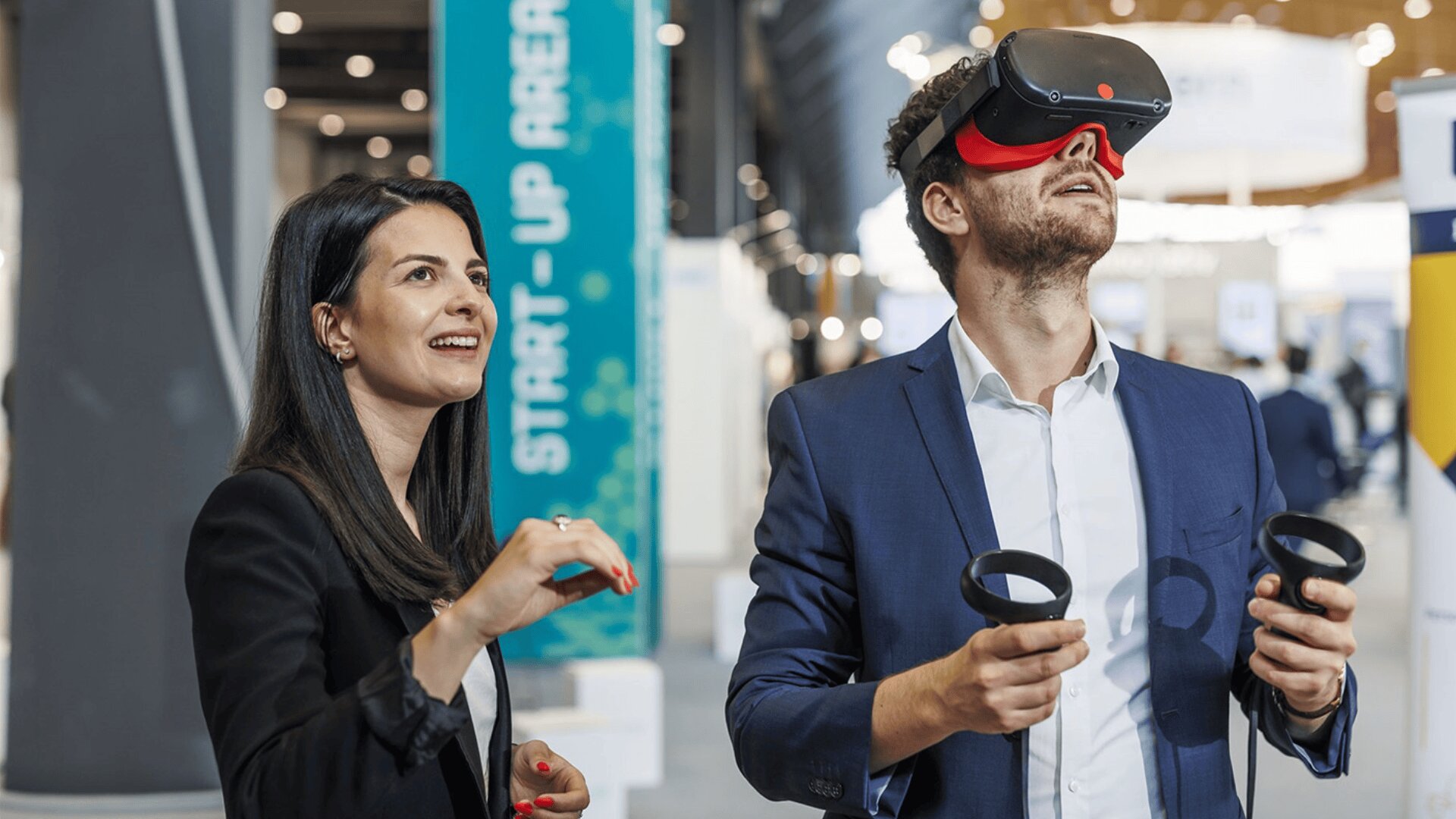 Eine Frau und ein Mann in Geschäftskleidung interagieren mit Virtual-Reality-Geräten; der Mann trägt eine VR-Brille und hält die Controller, während die Frau lächelt und gestikuliert, als sei sie in das Erlebnis vertieft.