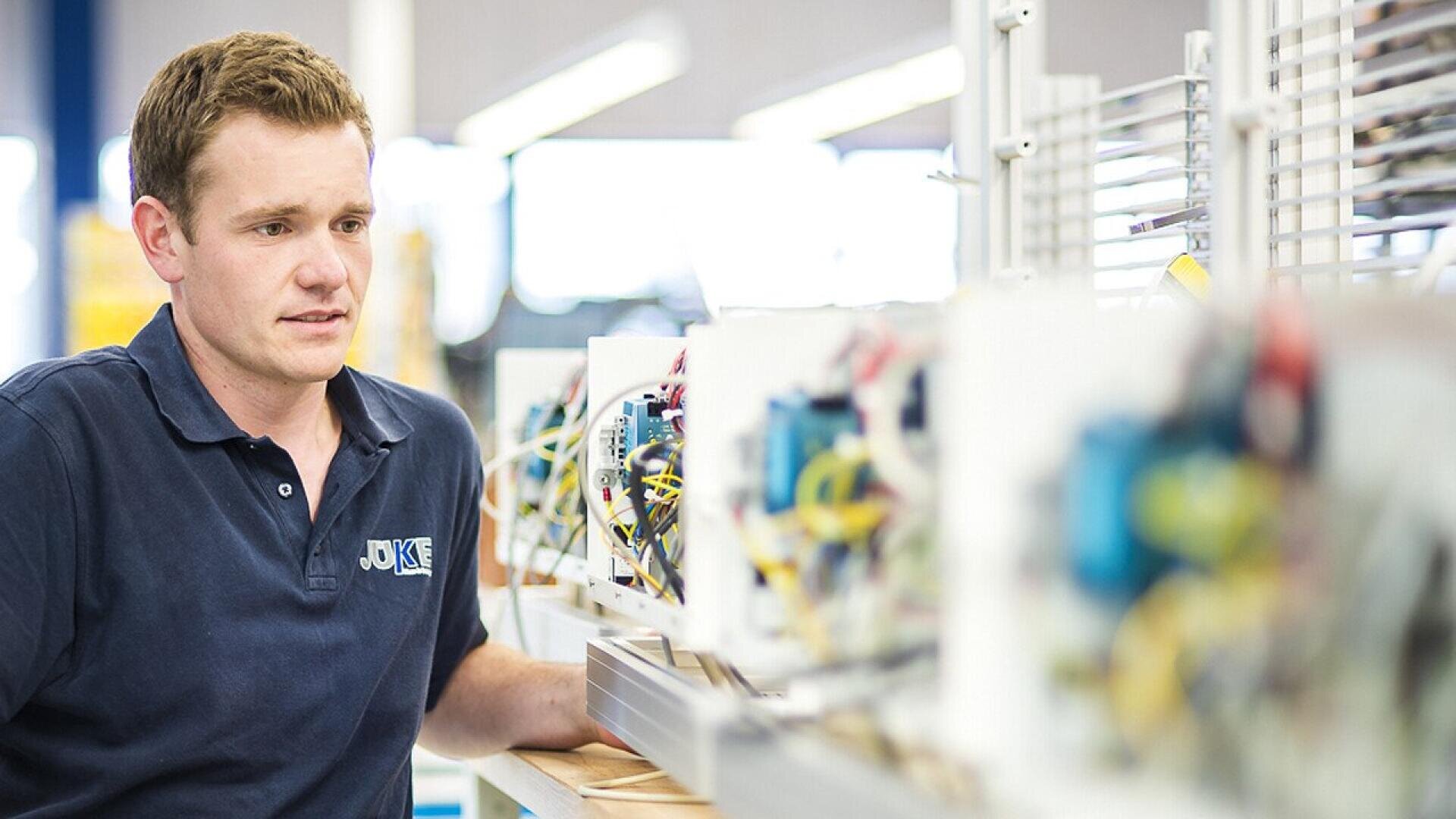 A man in a navy blue shirt stands beside a row of electronic equipment with exposed wiring in a brightly lit workshop or laboratory.