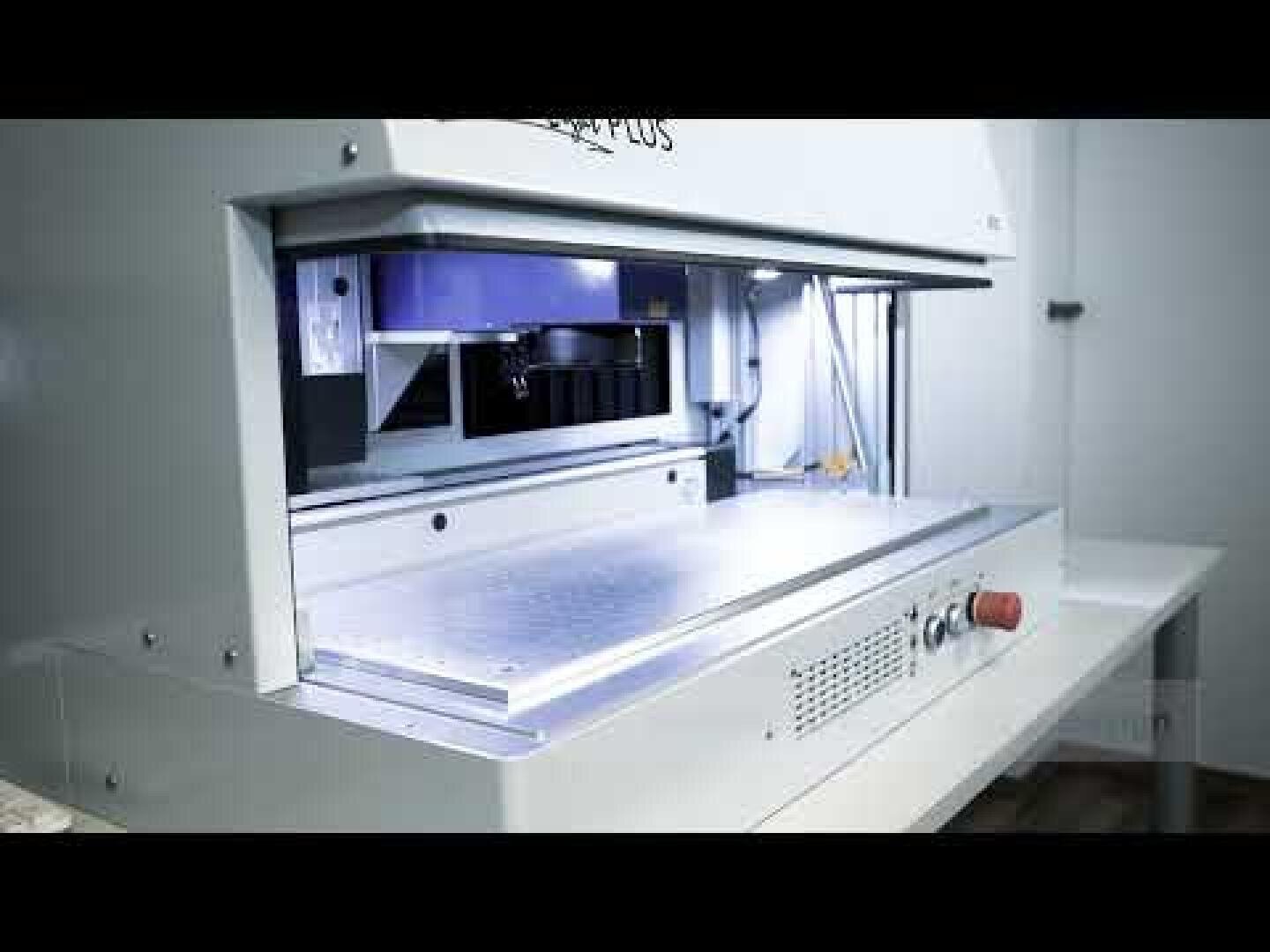 A close-up view of an industrial cutting or engraving machine in a well-lit room, featuring a metallic flat surface, control buttons, and a partially open work area.