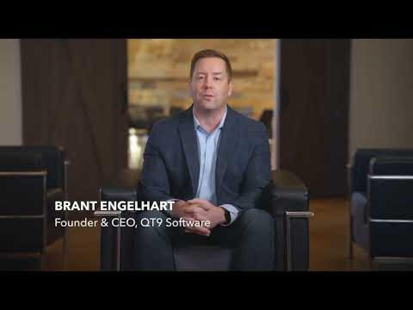 Ein Mann im Anzug sitzt auf einem schwarzen Stuhl in einer Büroumgebung. Der Text auf dem Bildschirm lautet: Brant Engelhart, Gründer & CEO, QT9 Software.