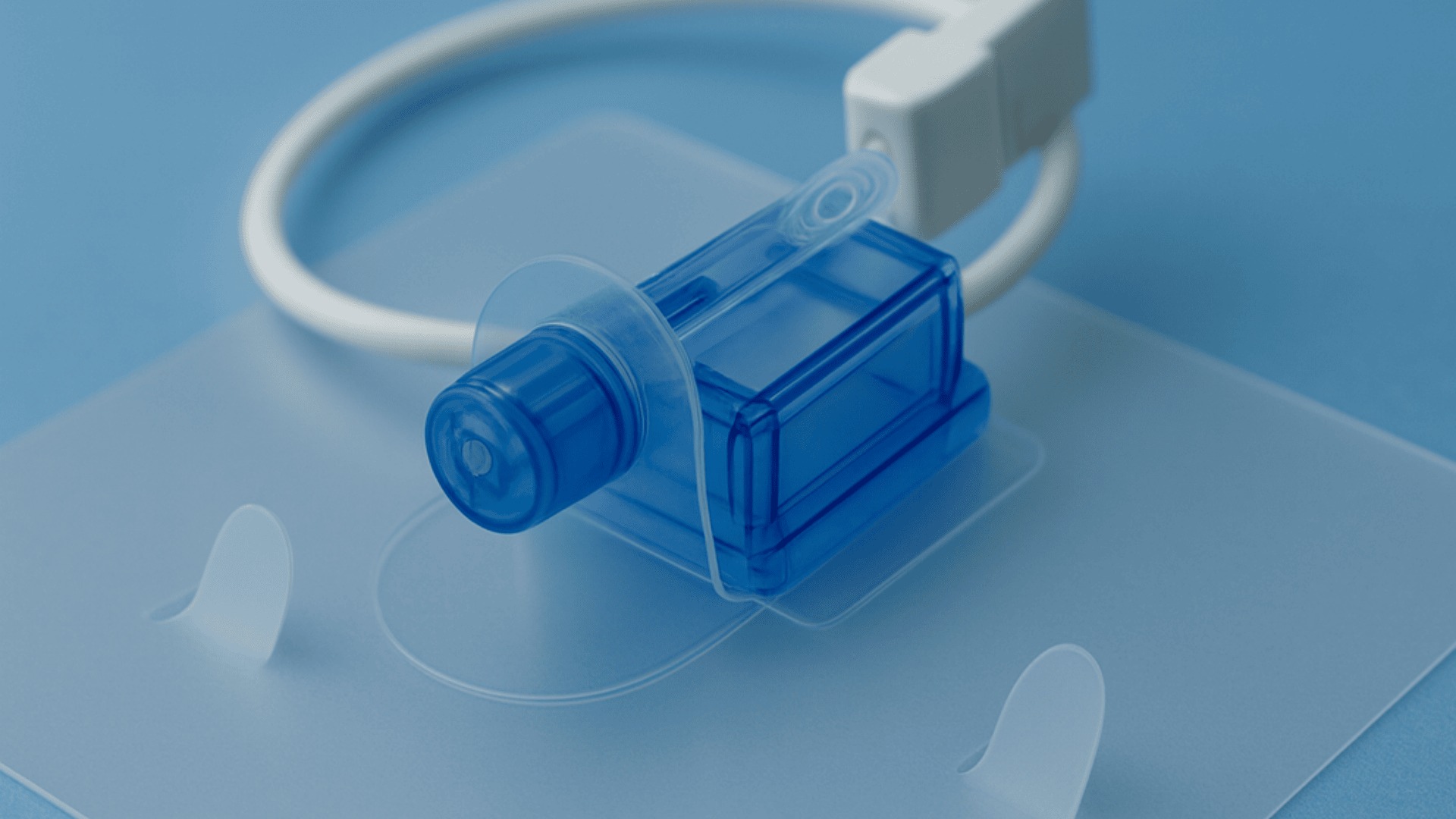 Close-up of a blue plastic medical plug attached to a clear adhesive patch, with a white cable in a loop in the background, against a light blue surface.