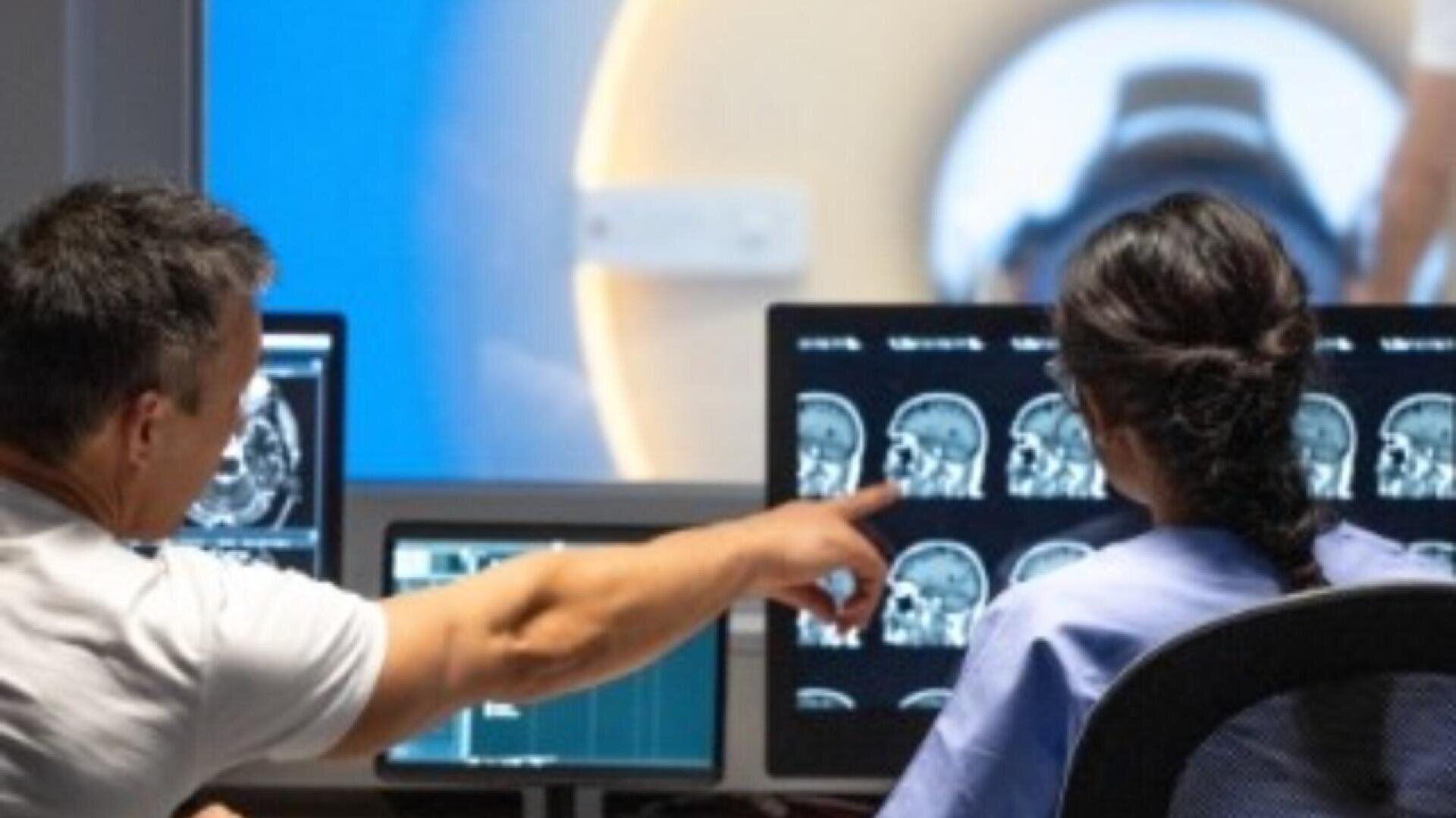 Two medical professionals review brain scan images on computer monitors, with one person pointing at the screens. An MRI machine is visible in the background.