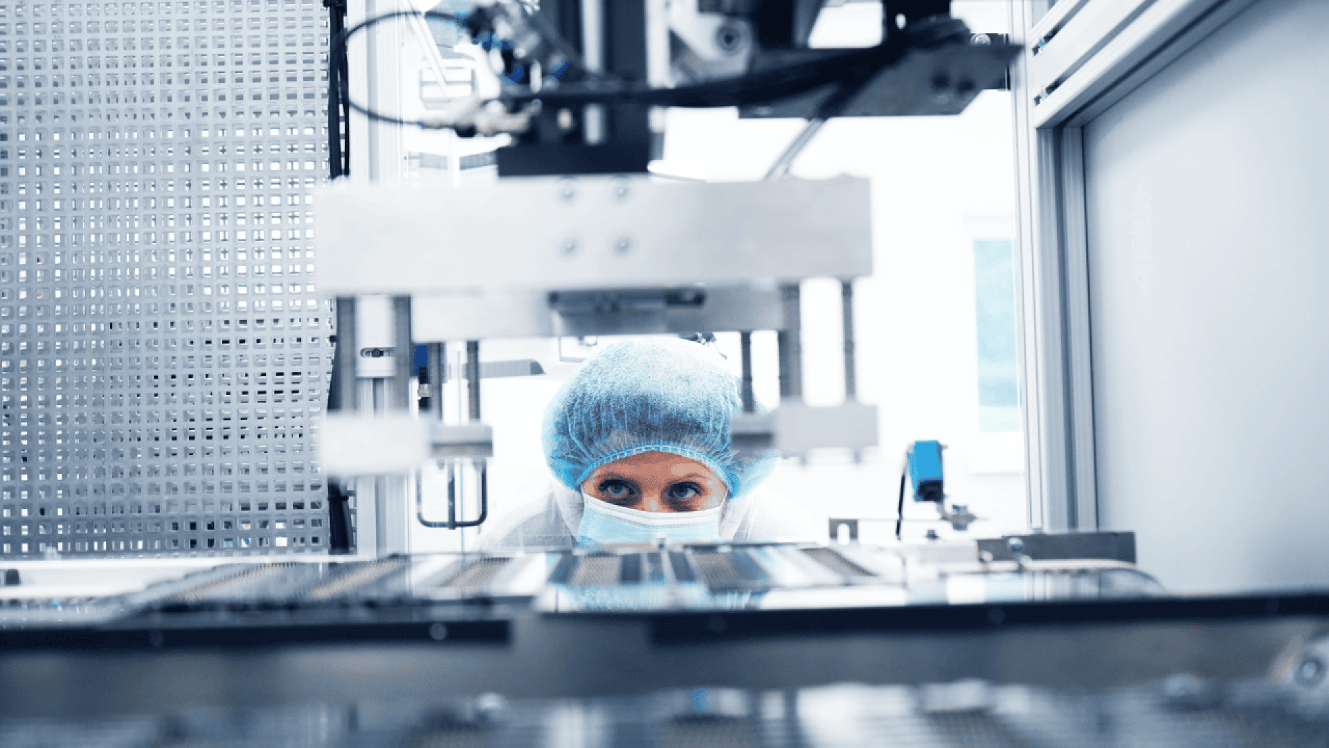 A person wearing a blue hair cover, face mask, and lab coat closely observes machinery in a sterile laboratory or manufacturing environment.