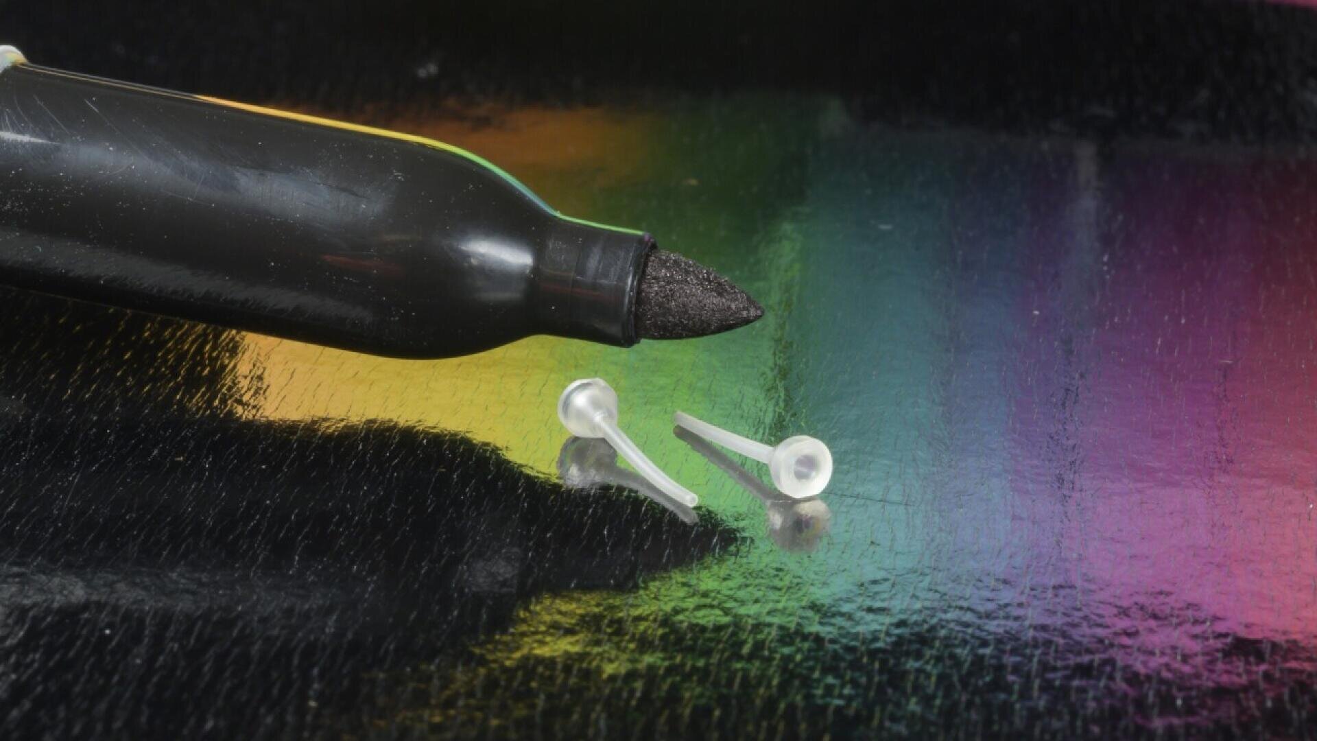 Eine Nahaufnahme eines schwarzen Markers, dessen Spitze neben zwei kleinen, durchsichtigen Ohrringverschlüssen aus Kunststoff auf einer glänzenden, mehrfarbigen Oberfläche sichtbar ist.