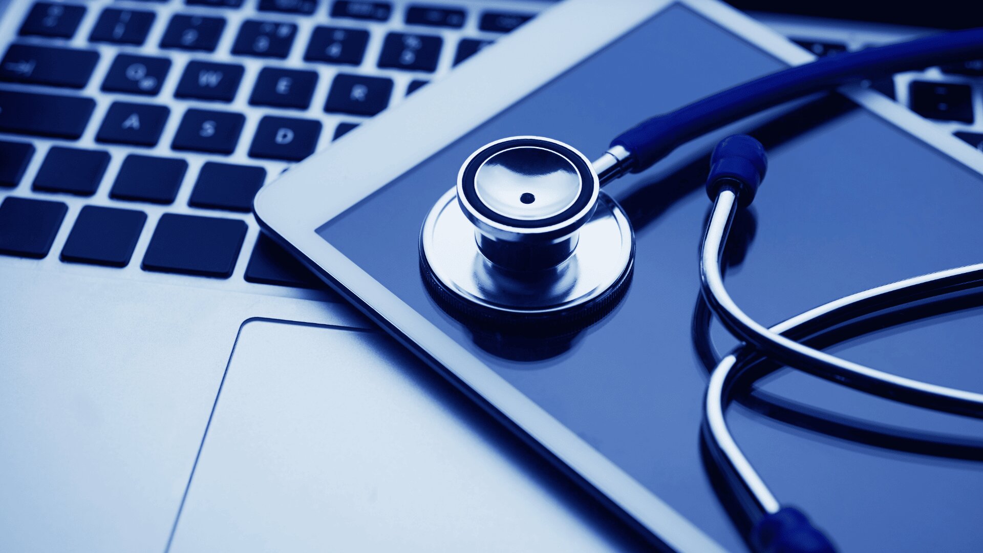Ein Stethoskop liegt auf einem Tablet, das wiederum auf einer Laptop-Tastatur platziert ist und die Schnittstelle zwischen Gesundheitswesen und Technologie symbolisiert.