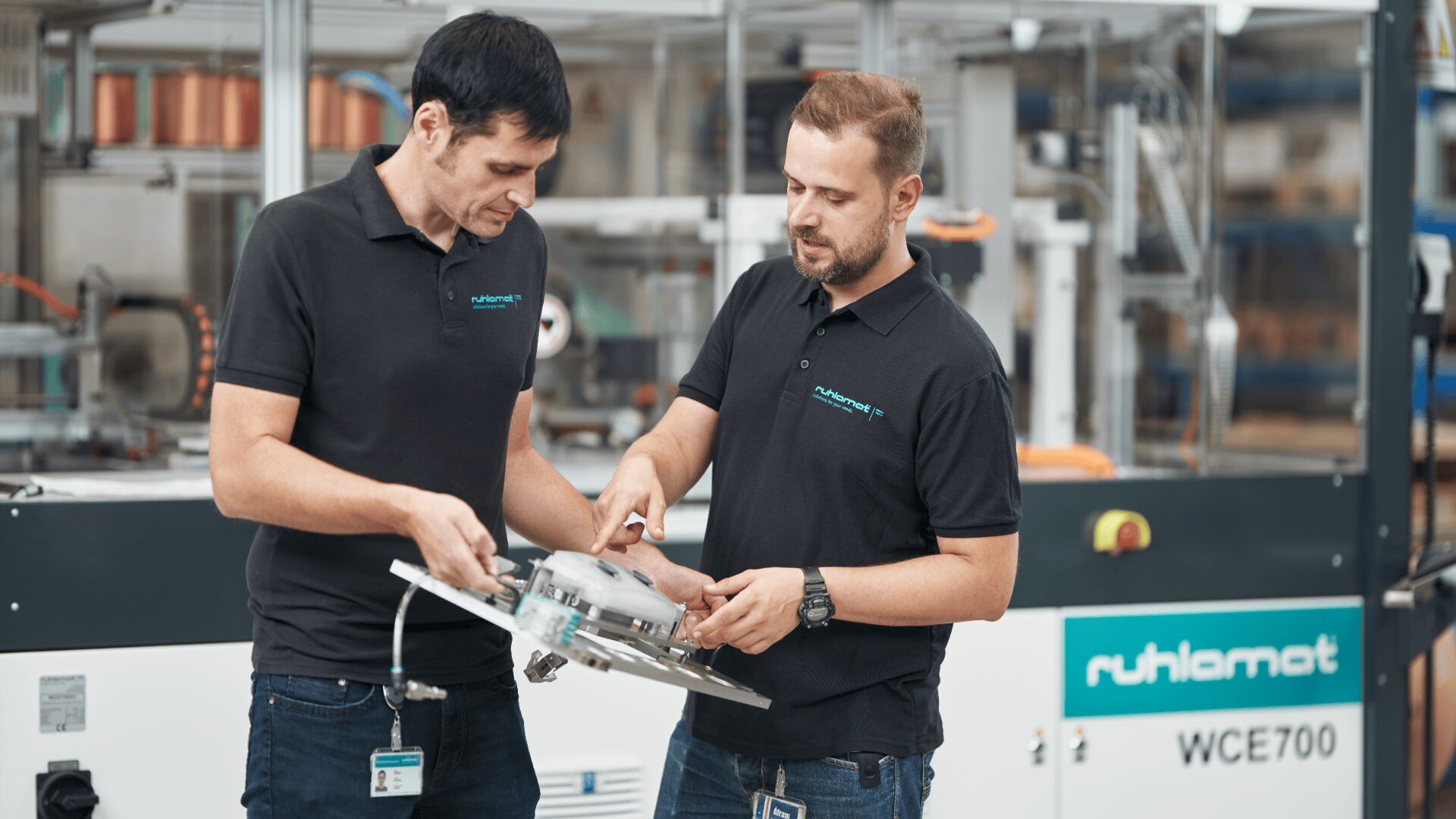 Zwei Männer in schwarzen Poloshirts untersuchen ein metallisches, rundes Maschinenteil in einer modernen Industrieumgebung. Sie stehen vor einem Gerät mit der Aufschrift „Ruthlamat WCE700“.