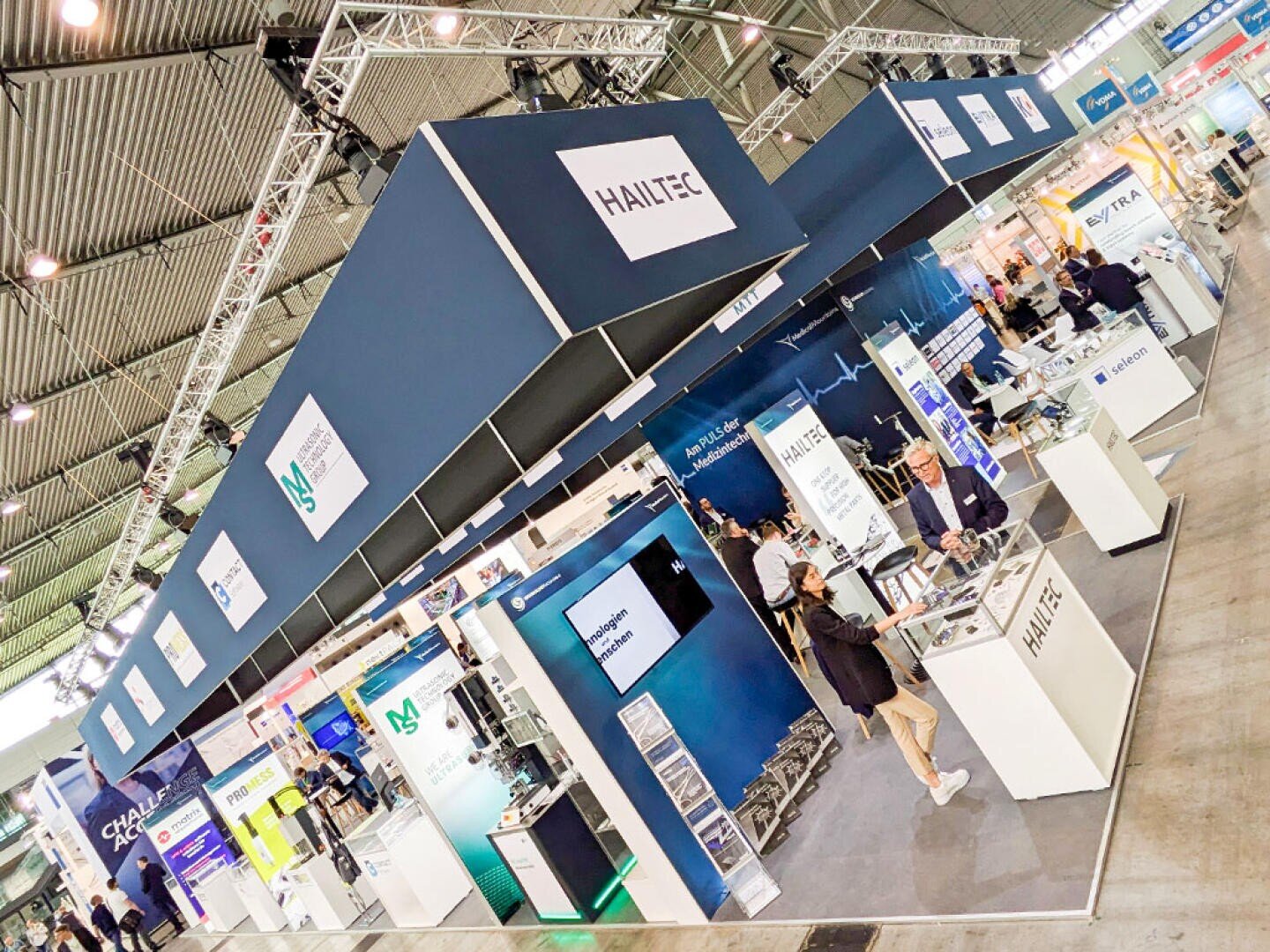 Ein großer Messestand mit MedicalMountains GmbH Branding zeigt Bildschirme, Theken und interagierende Menschen. Der Stand ist Teil einer belebten Messehalle mit anderen Firmenständen und Bannern im Hintergrund.