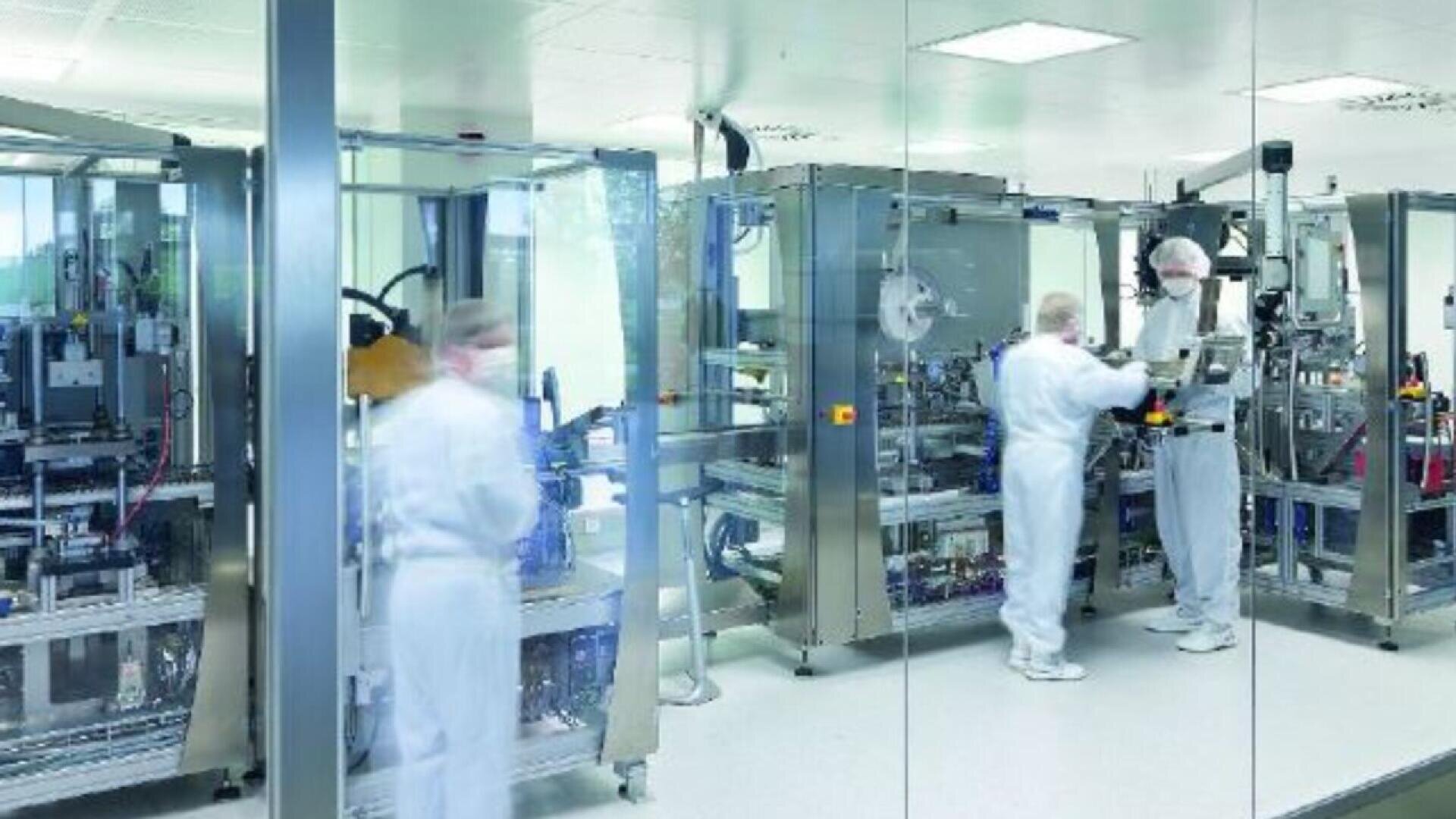 Three people in white protective suits and hair covers work with large, high-tech machinery inside a clean, bright laboratory or manufacturing facility, seen through a glass wall.