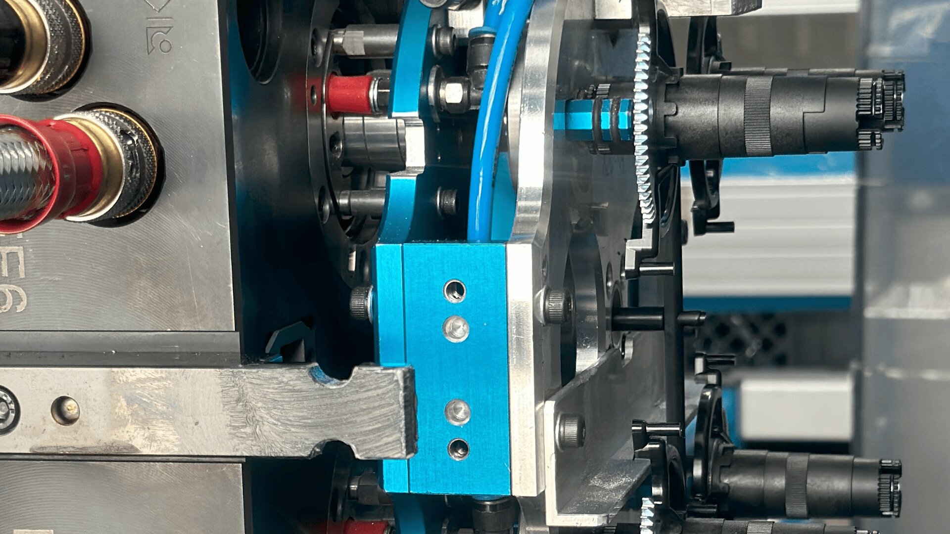 Nahaufnahme einer Industriemaschine mit blauen und silbernen Metallteilen, schwarzen Zahnrädern, Bolzen und roten Verbindungsstücken, die die komplexe Mechanik der Anlage verdeutlichen.