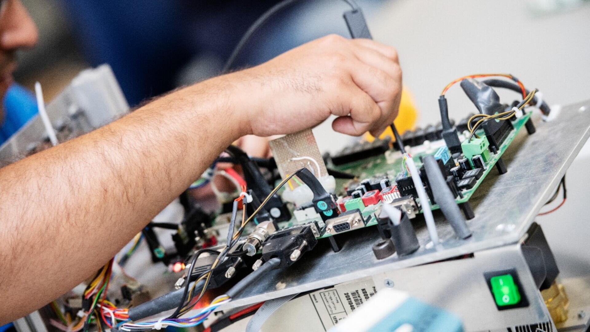 A person’s hand uses a tool to work on an open electronic circuit board with various wires and components visible.