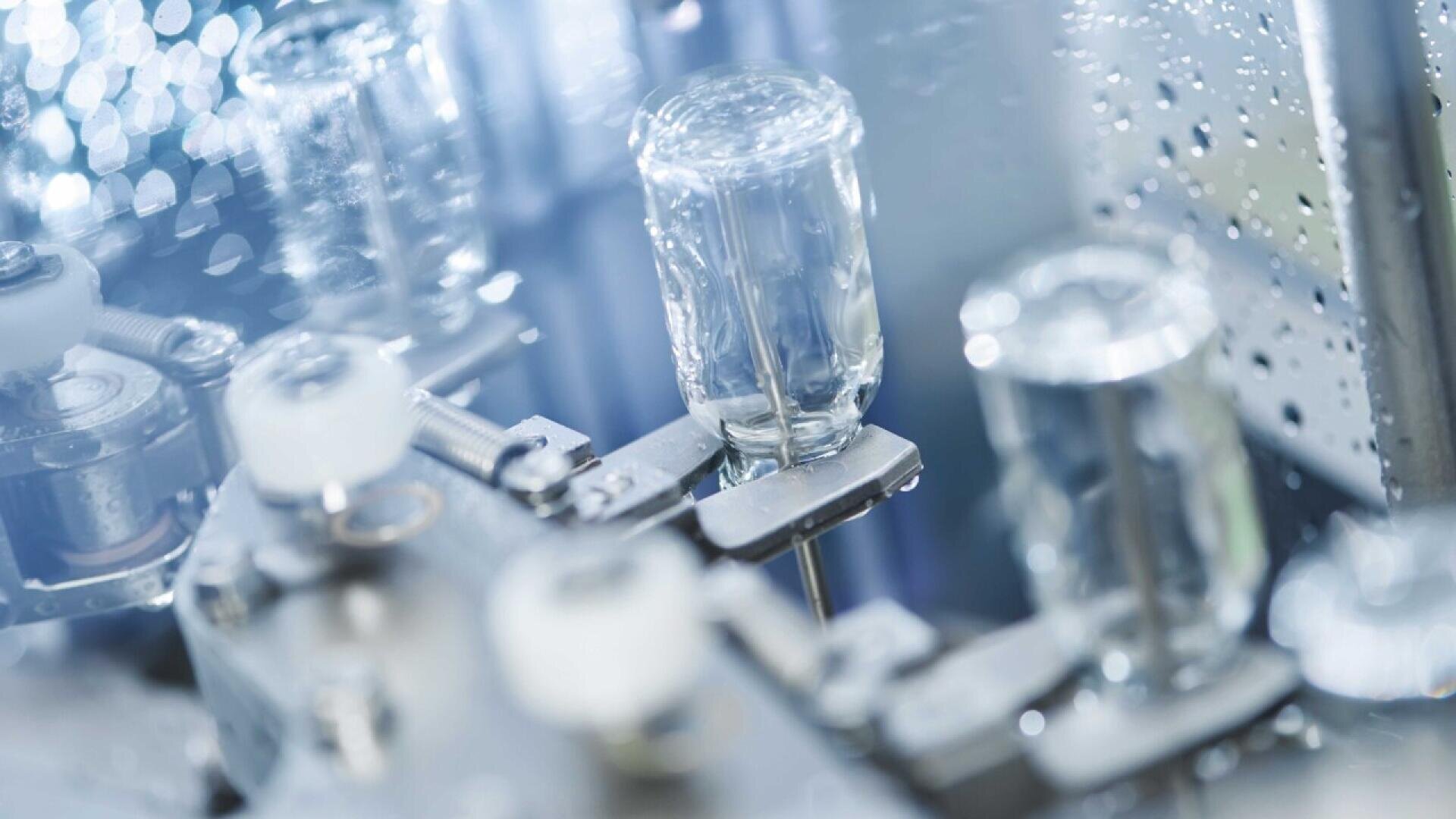Close-up of glass vials being filled or processed by automated machinery in a sterile, high-tech laboratory or pharmaceutical manufacturing environment.