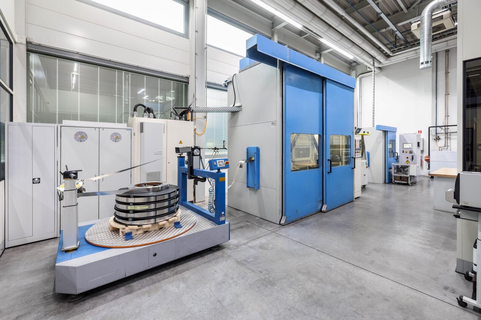 Eine moderne Industrieanlage mit großen blauen und weißen Maschinen, Metallgeräten und einer runden Plattform mit gestapelten Metallteilen, alles in einer sauberen, gut beleuchteten Umgebung mit Betonboden.