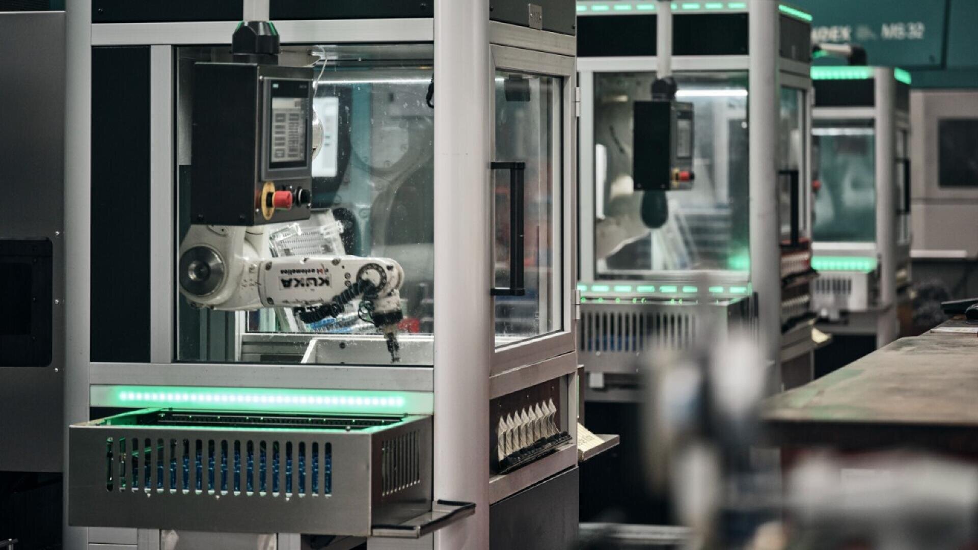 Robotic arms operate inside enclosed manufacturing stations, with illuminated control panels and safety glass, in a modern industrial facility.