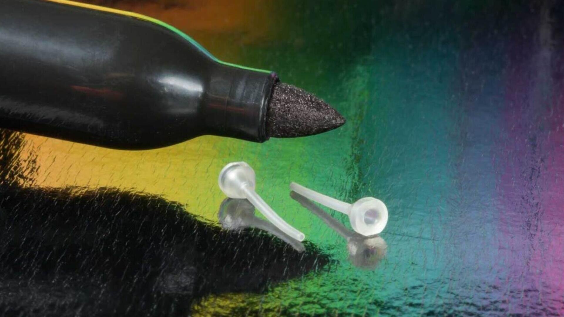 Eine Nahaufnahme einer schwarzen Markerspitze auf einer glänzenden, mehrfarbigen Oberfläche, neben zwei kleinen, durchsichtigen Plastikkappen.