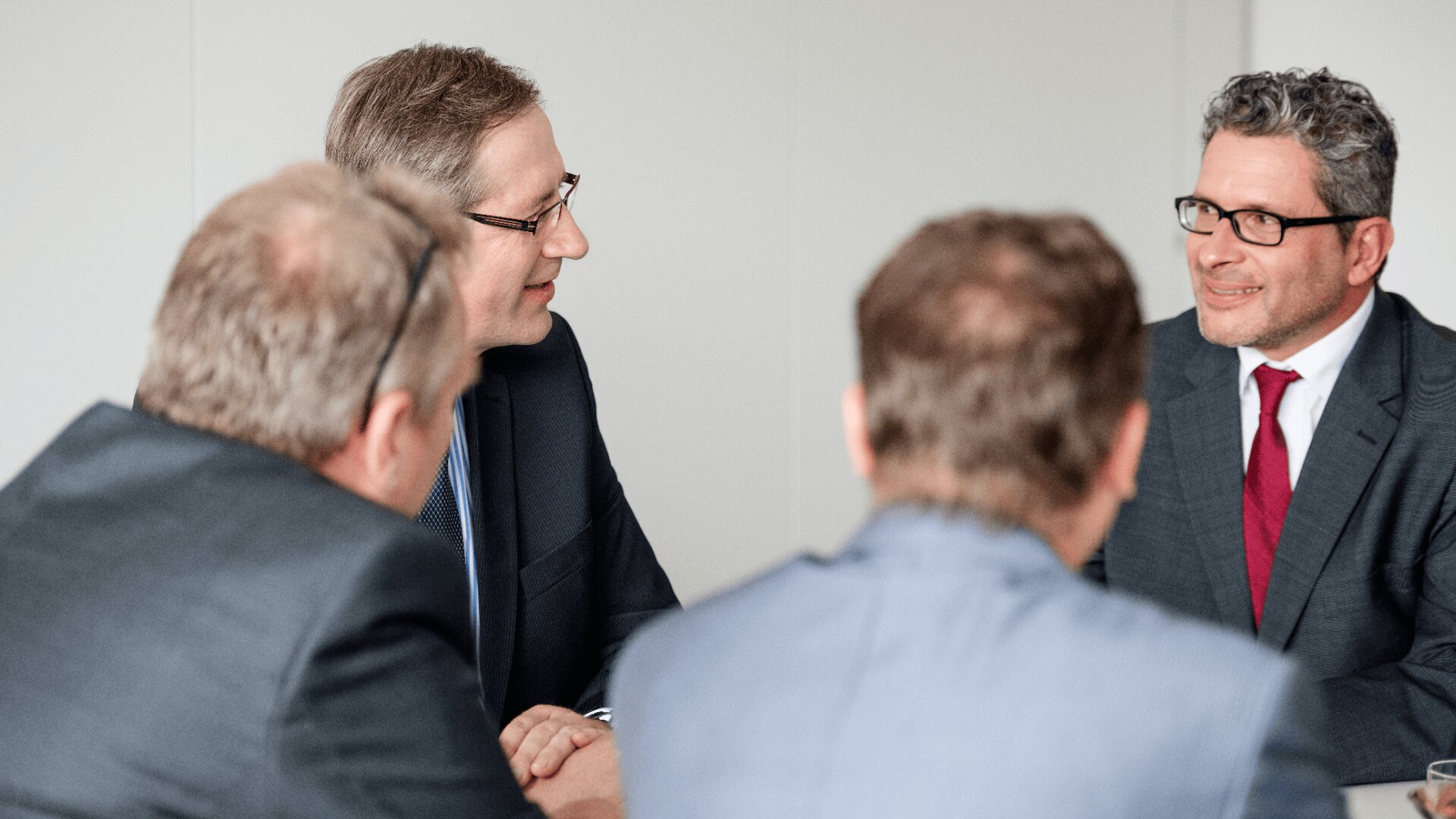 Vier Männer in Businessanzügen sitzen um einen Tisch und unterhalten sich. Zwei von ihnen blicken mit verschwommenen Gesichtern in die Kamera, während die anderen beiden lächelnd einander in einem hell erleuchteten Büro gegenüberstehen.