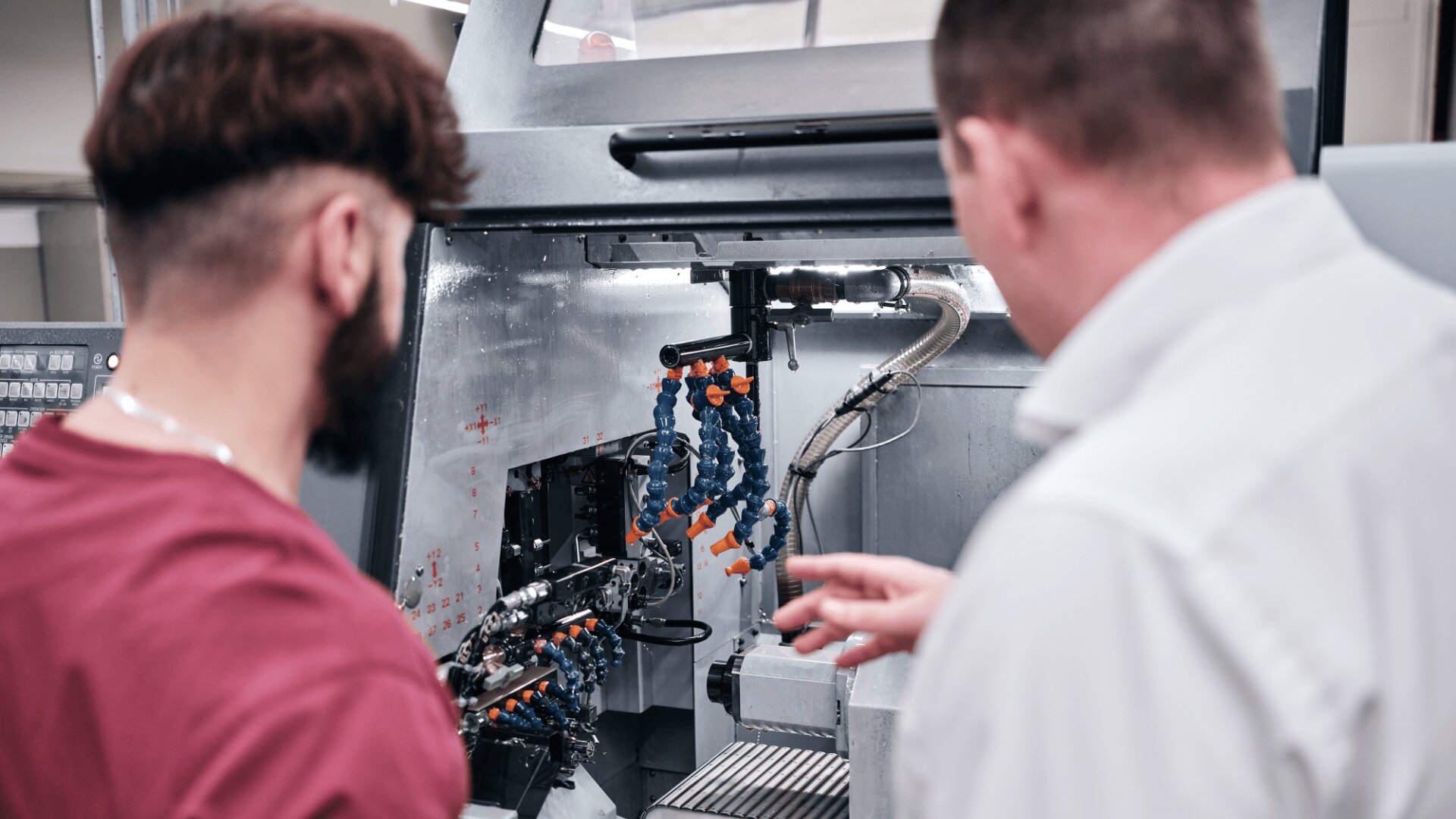 Zwei Männer stehen vor einer CNC-Maschine, prüfen ihre Innenkomponenten und besprechen ihre Funktionsweise. Einer trägt ein rotes Hemd, der andere ein weißes Hemd.