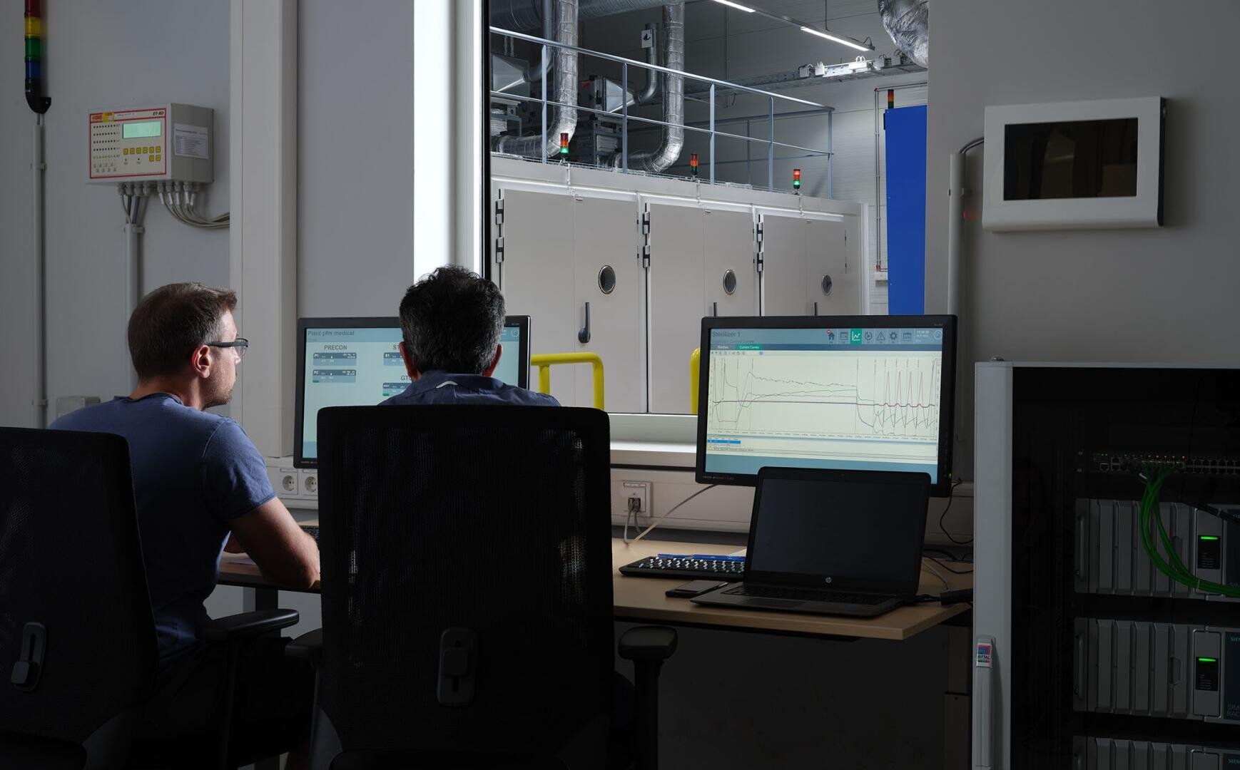 Zwei Personen sitzen an Schreibtischen und überwachen Daten und Wellenformen auf Computerbildschirmen in einem Kontrollraum. Durch ein großes Fenster sind Industrieanlagen und Rohrleitungen in einem hell erleuchteten Arbeitsbereich dahinter zu sehen.