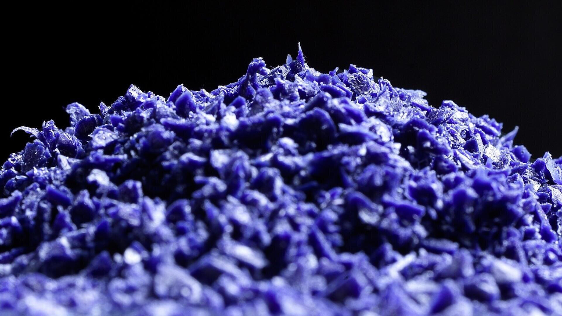 A close-up of a pile of vivid blue, shredded or flaked material, possibly plastic or wax, with a dark background. The texture is rough and jagged, highlighting the individual fragments.