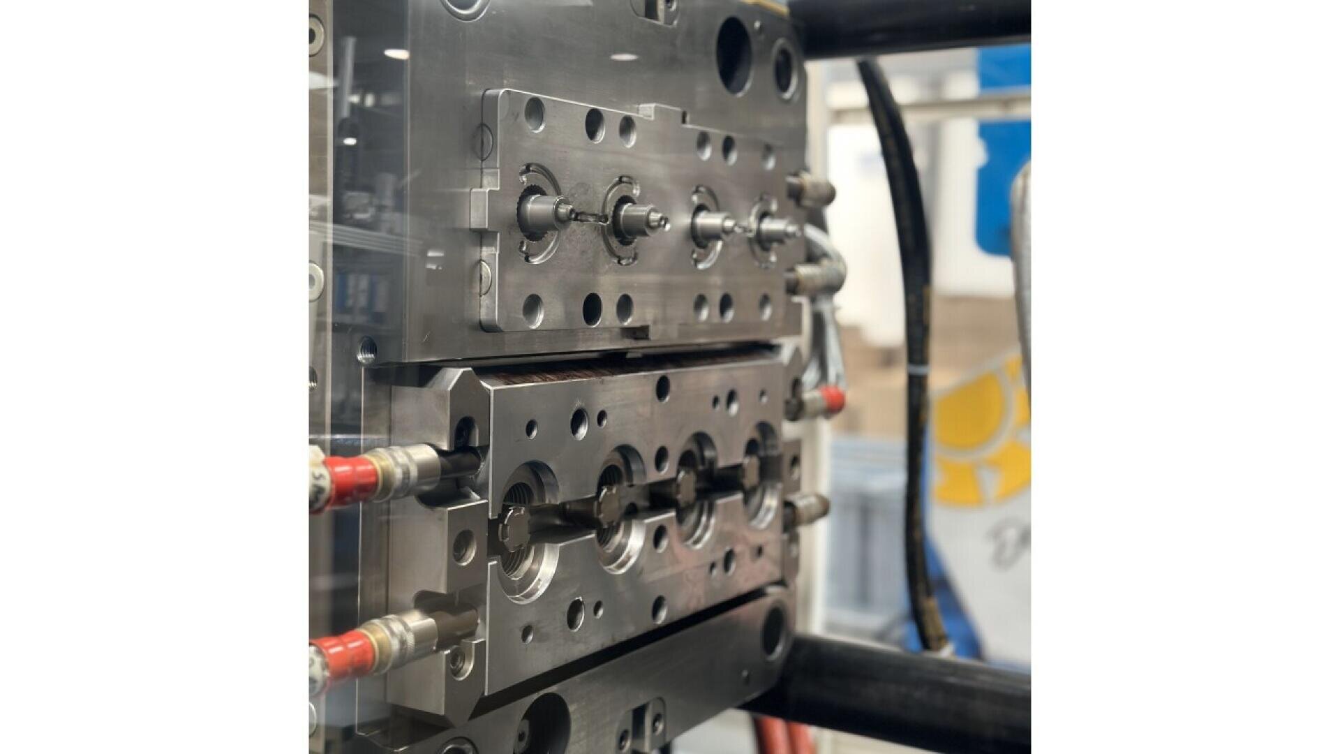 Close-up of a metal injection mold tool in a manufacturing setting, showing detailed engraved parts and multiple cylindrical components, with red hoses and industrial equipment visible in the background.