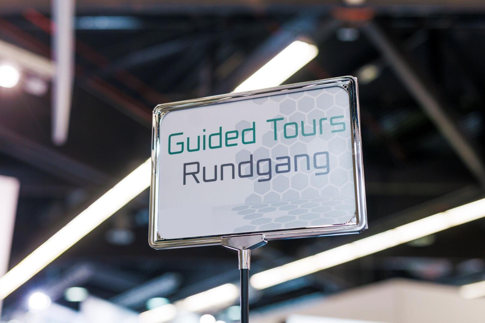 Ein Schild mit der Aufschrift Guided Tours Rundgang in grüner und schwarzer Schrift, mit einem Sechseckmuster im Hintergrund. Das Schild wird in einem Innenraum hochgehalten, wahrscheinlich bei einer Veranstaltung oder Ausstellung, wobei dahinter verschwommene Lichter und Deckenbalken zu sehen sind.