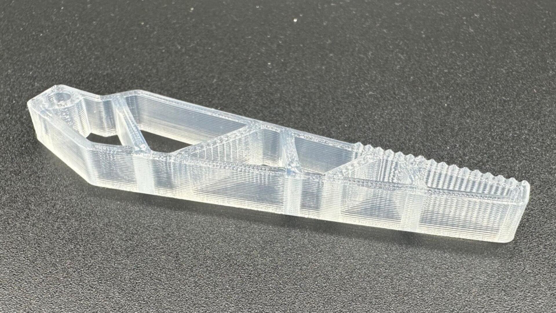 A translucent, 3D-printed plastic tool with an angular design, featuring triangular cutouts and a textured, ridged end lying on a black surface.