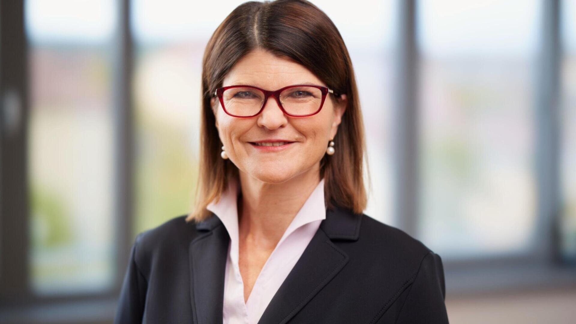 A woman with straight brown hair, wearing red glasses, pearl earrings, and a dark blazer over a light blouse, smiles in front of a blurred office window background.