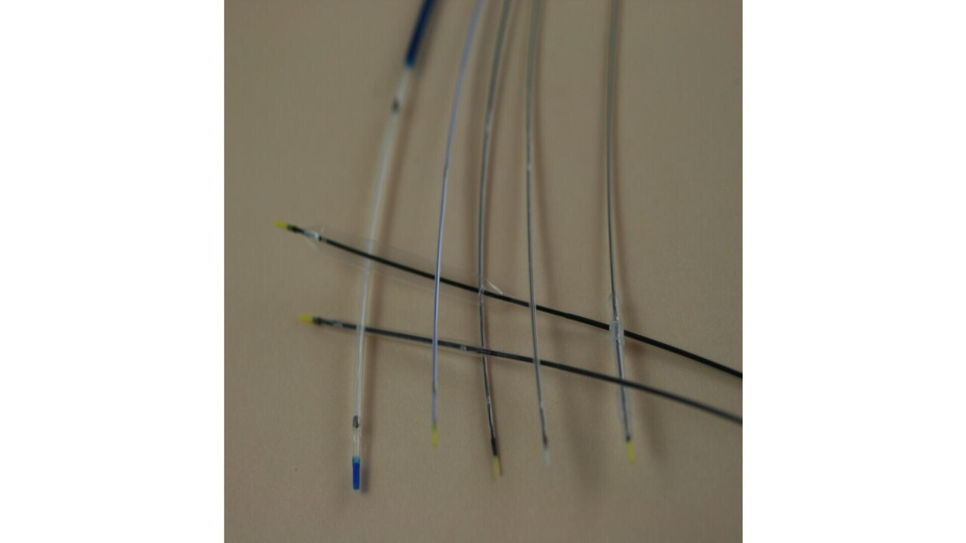 Several thin, flexible medical wires and catheters with colored tips, mostly yellow and blue, are arranged parallel on a neutral background.