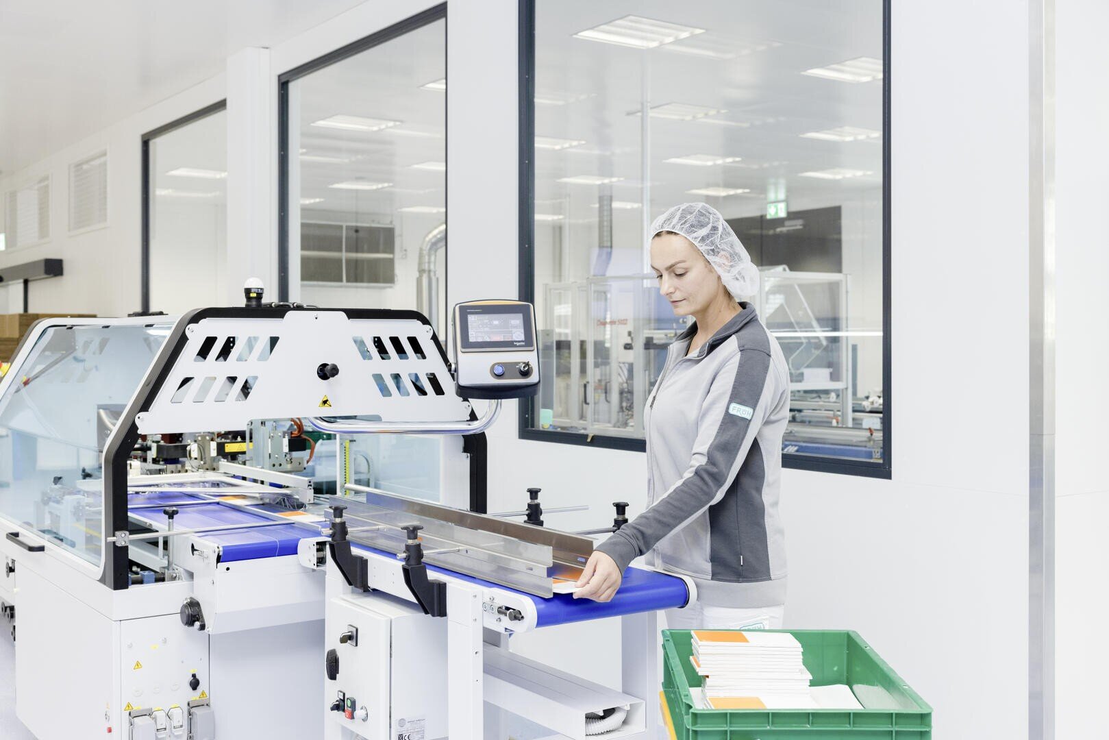 A worker in a hair net and uniform operates a machine on a production line in a clean, bright factory, with stacks of paper or packaging material in a green bin nearby.