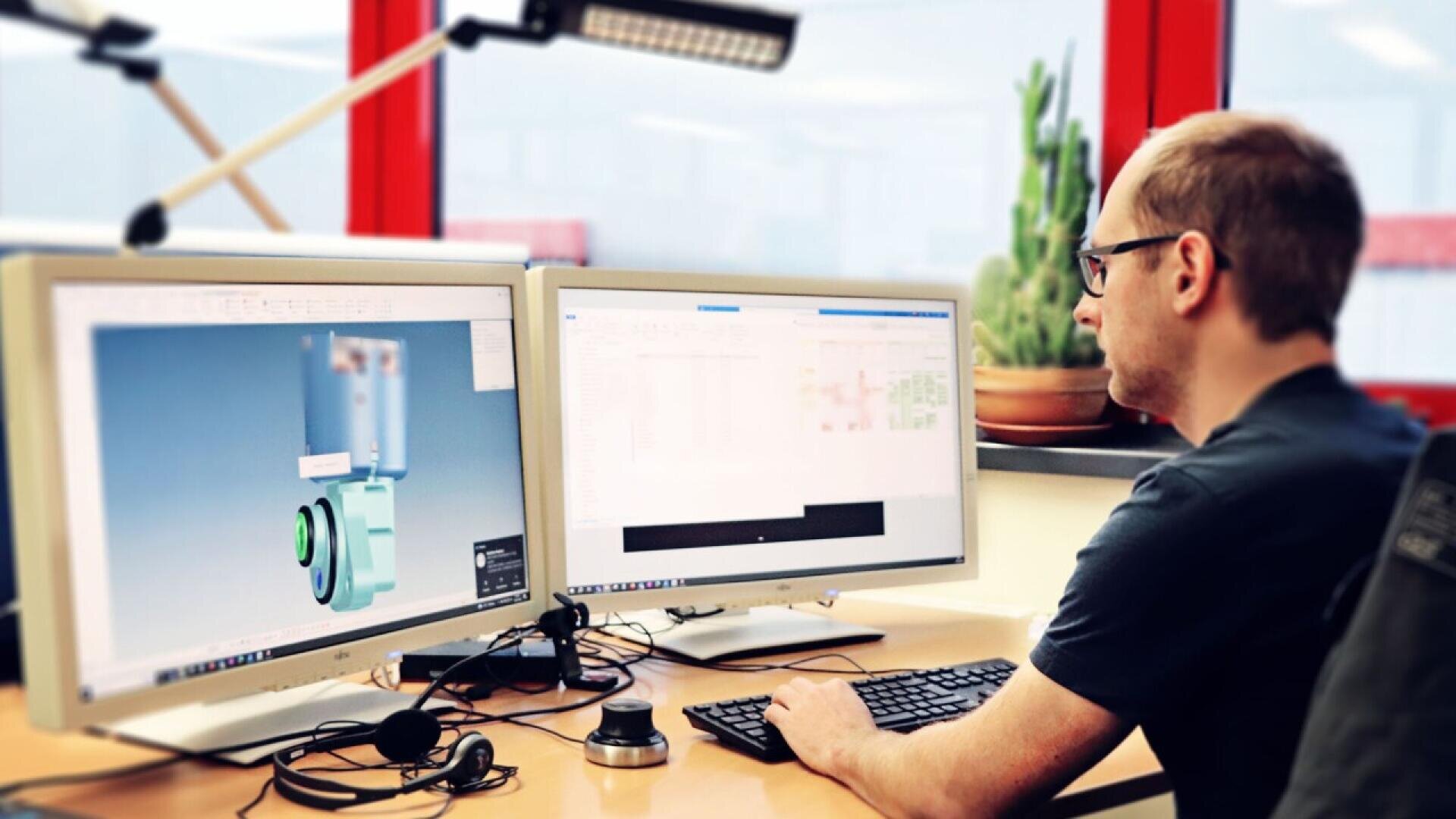 A person sits at a desk working at two computer monitors; one shows a 3D model while the other displays code or data. A keyboard, mouse, and headset are on the desk, with a plant and window in the background.