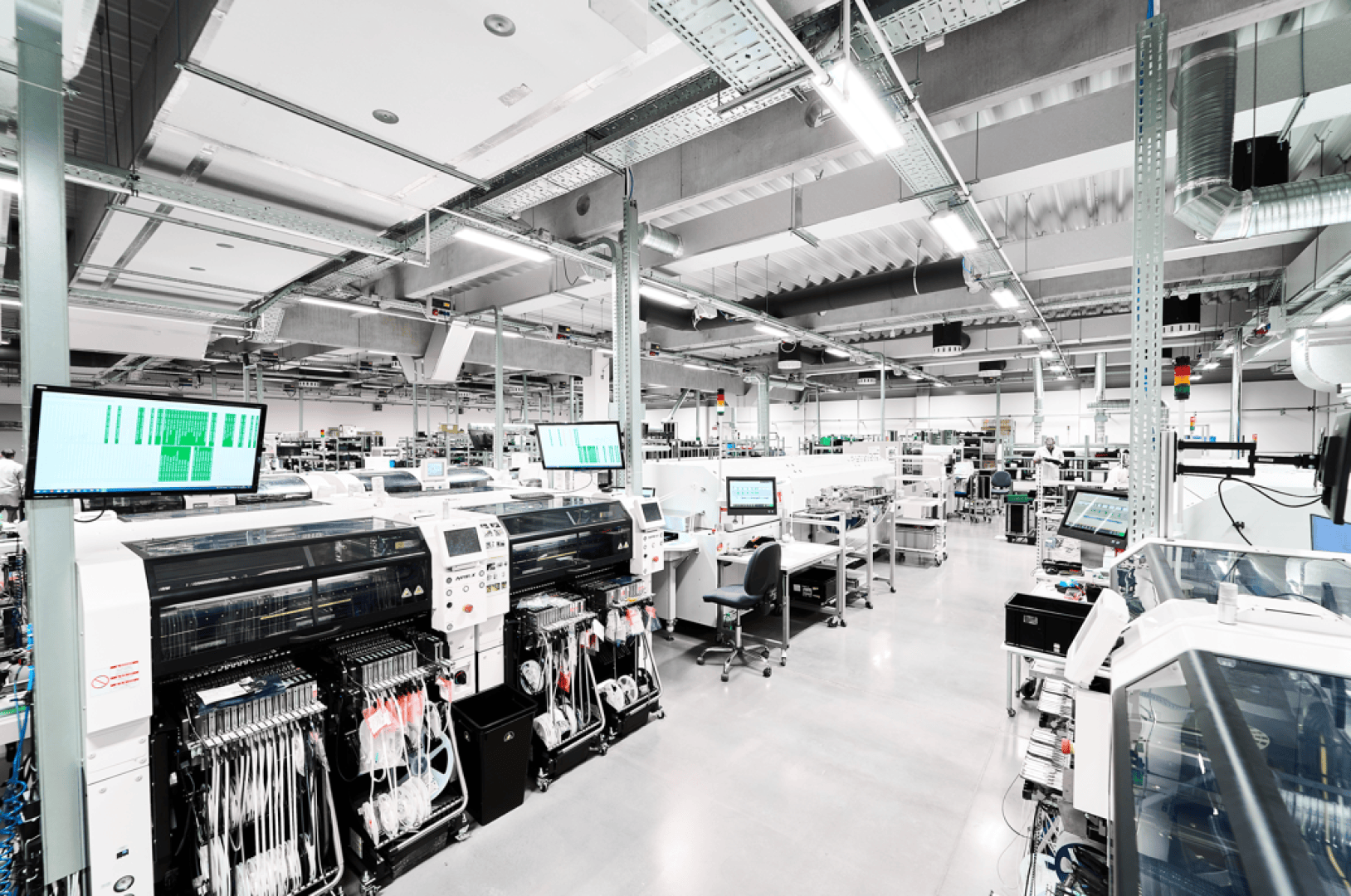 A modern electronics manufacturing facility with automated machines, conveyor systems, workstations and multiple computer monitors for displaying data, all in a bright, organized industrial space.
