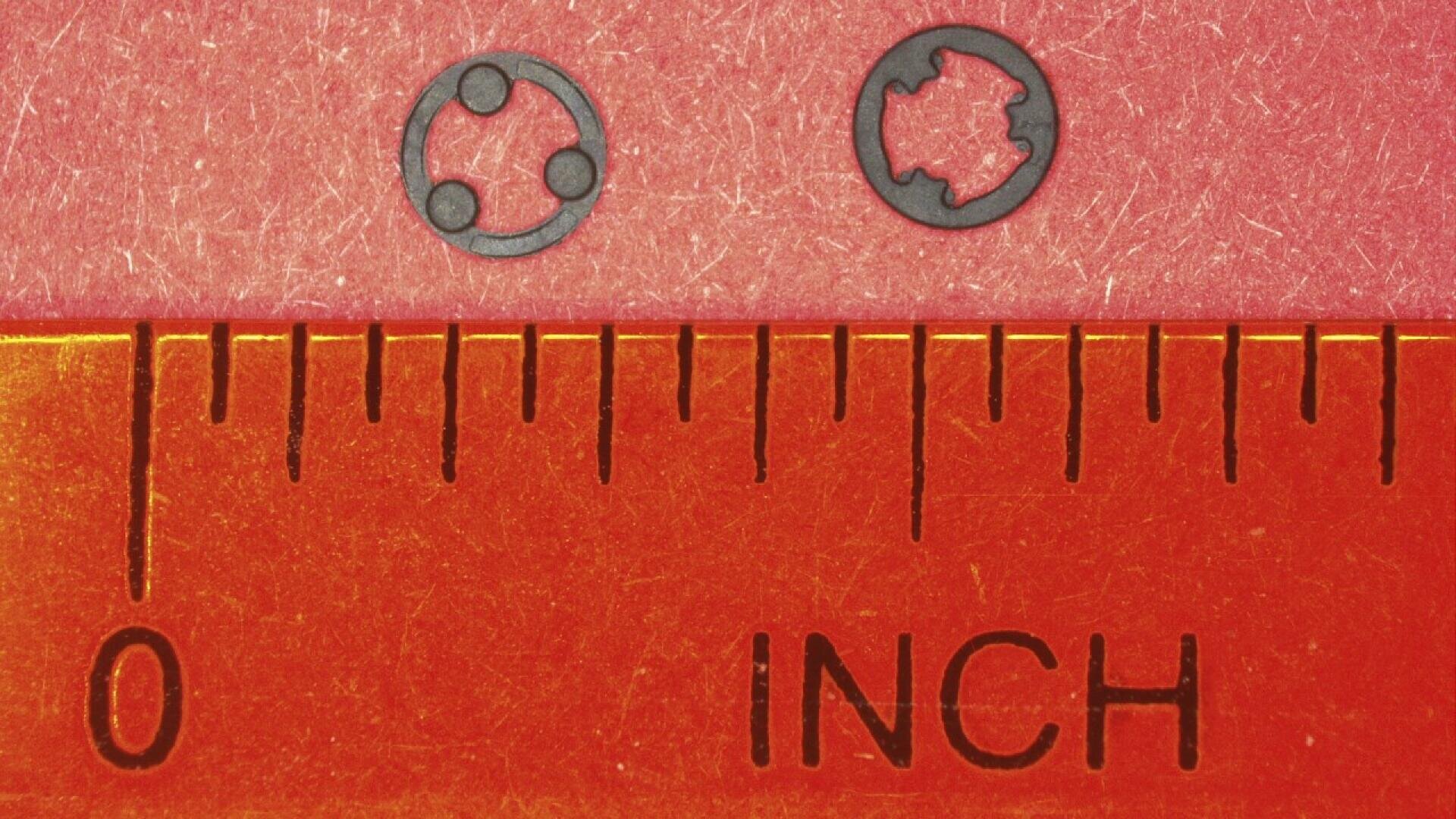 Two tiny circular metal washers with different inner designs are placed above an orange ruler, which shows inch measurements starting from zero, highlighting their small size.