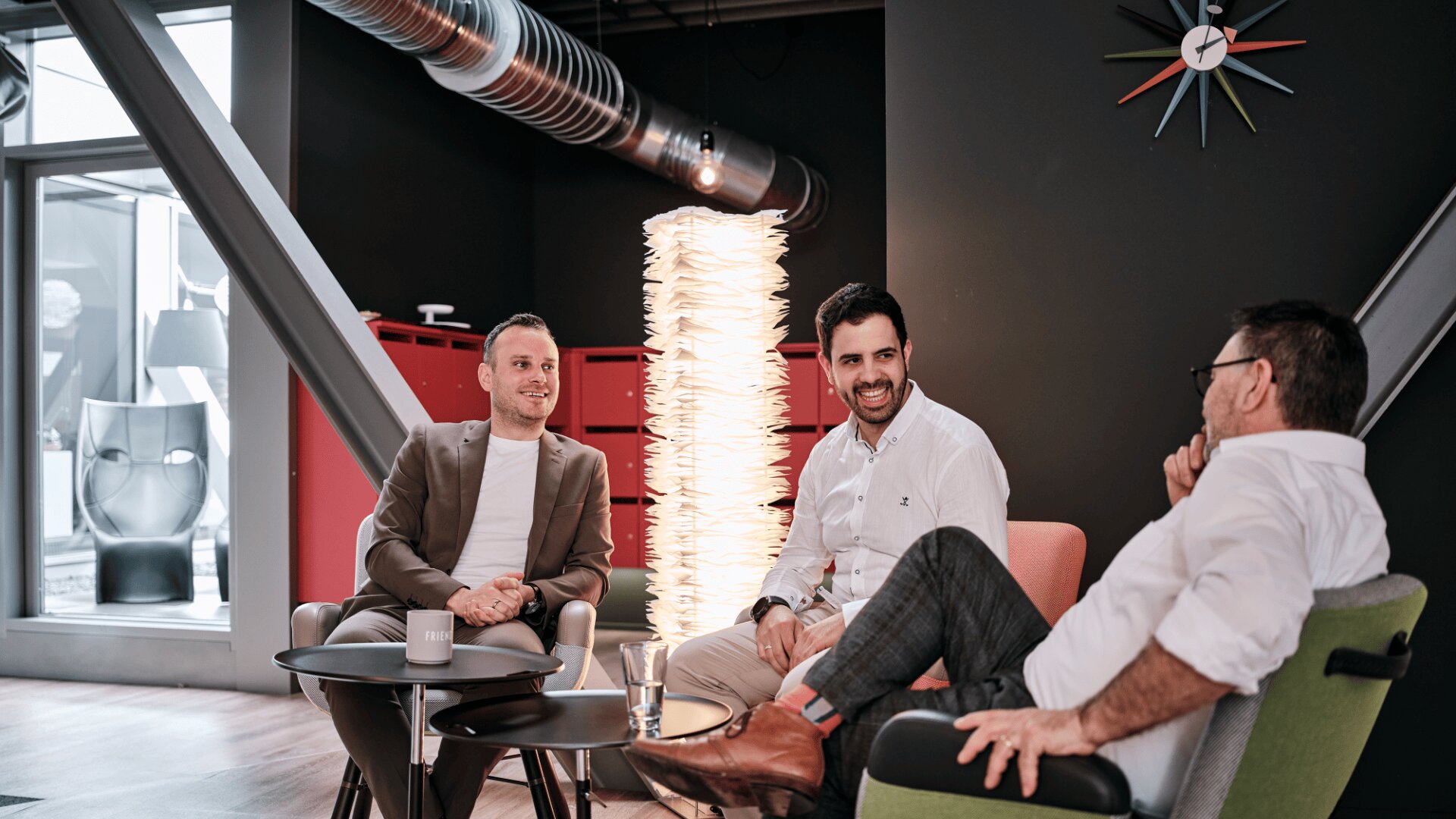 Drei Männer in legerer Business-Kleidung sitzen lächelnd und unterhalten sich auf modernen Stühlen in einer stilvollen Bürolounge mit einer einzigartigen Stehlampe, einer Wanduhr und roten Schränken im Hintergrund.