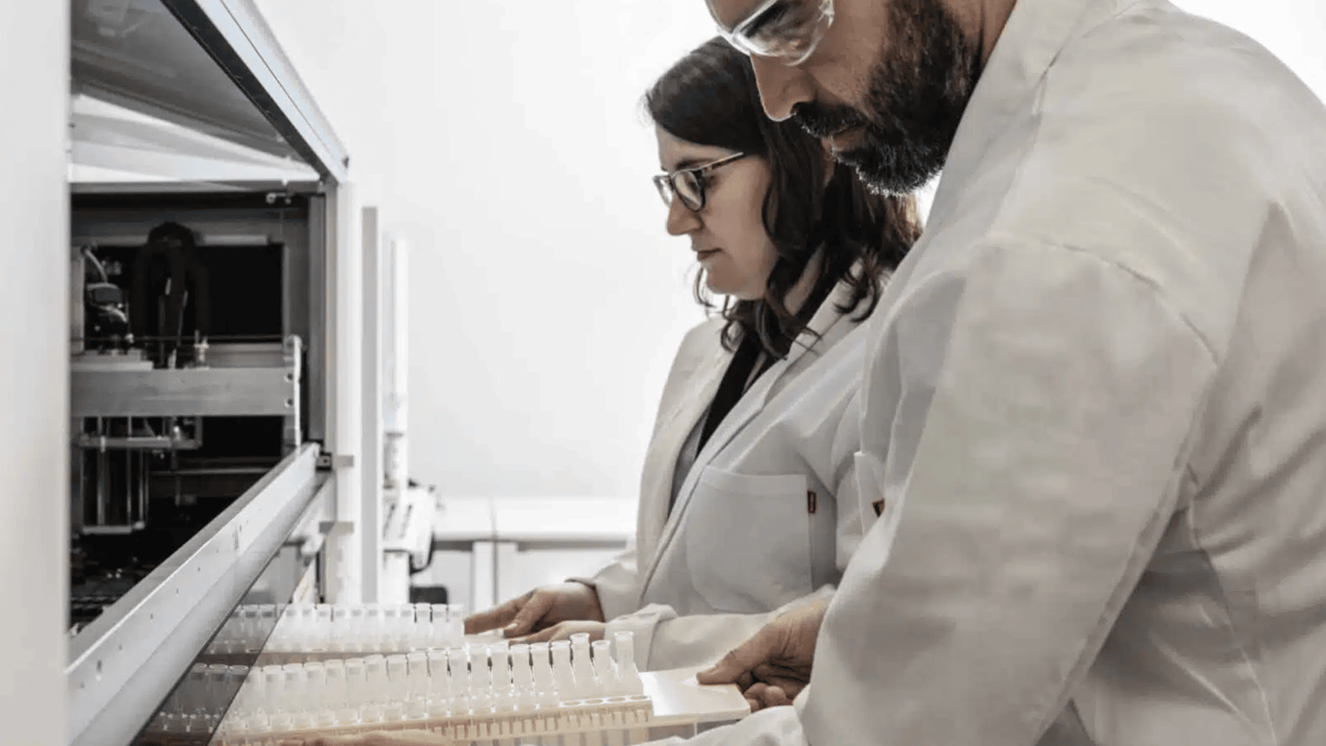 Two scientists wearing lab coats and safety glasses work with test tubes and laboratory equipment in a modern lab setting.