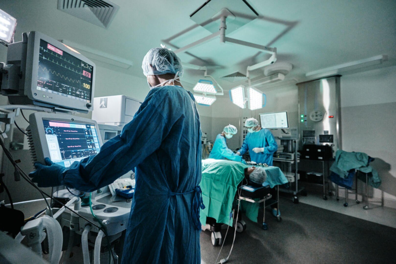 Medizinisches Fachpersonal in OP-Kitteln und Masken führt in einem hell erleuchteten Operationssaal eine Operation durch. Eine Person überwacht die Vitalwerte auf einem Bildschirm, während andere einen Patienten auf dem OP-Tisch behandeln.