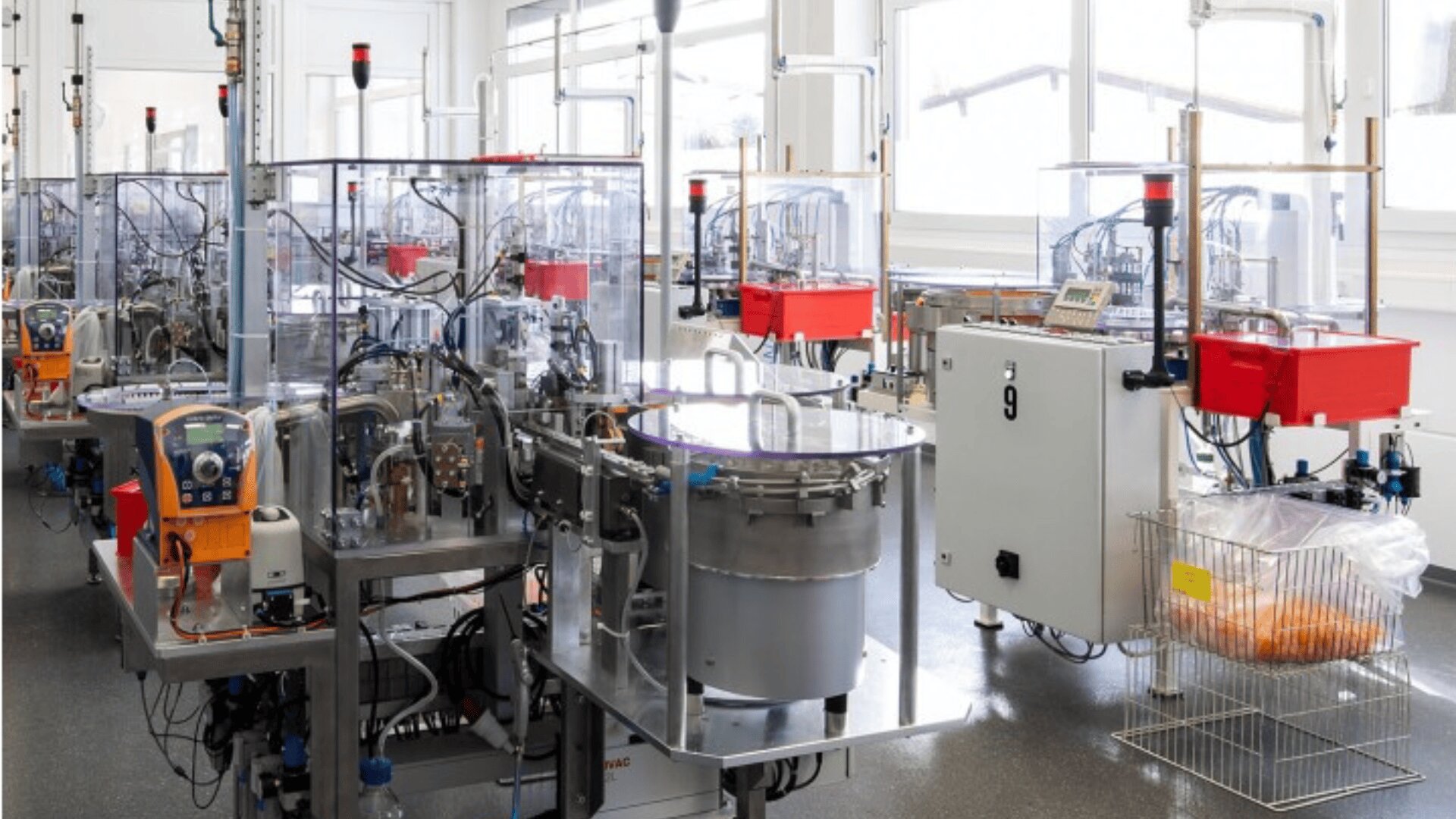 A laboratory room filled with automated laboratory equipment, machines with transparent covers, control panels, and a wire rack holding bags, all under bright window lighting.